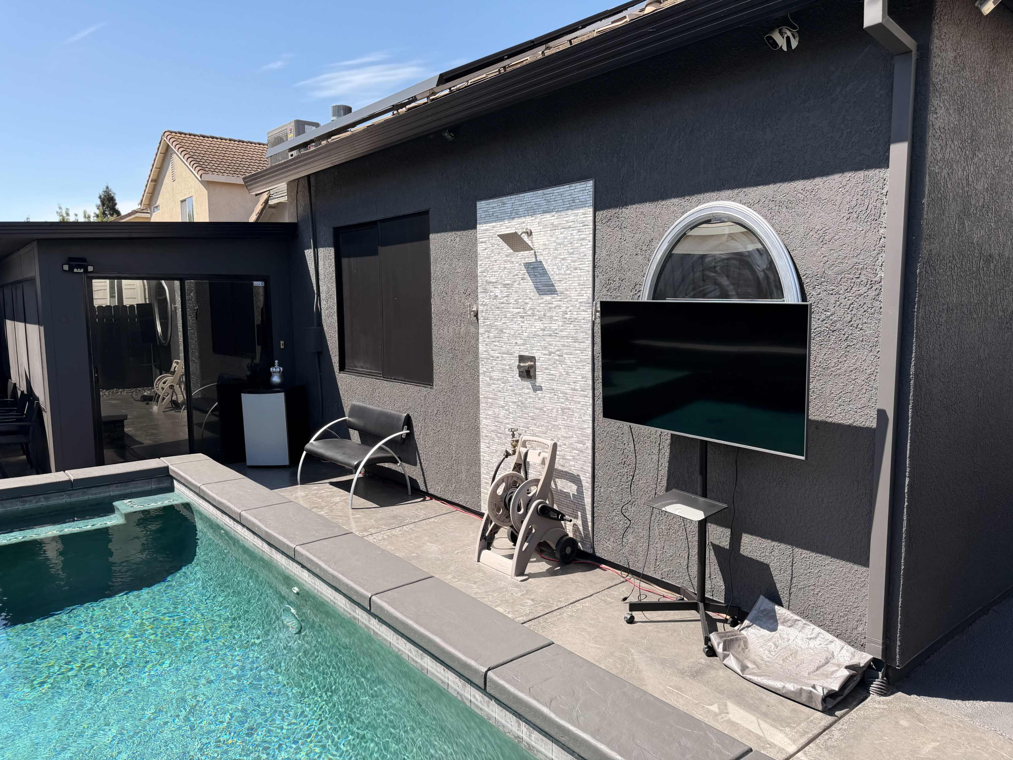 The image shows a modern backyard with a swimming pool, a gray exterior wall featuring a round window, and a mounted television.