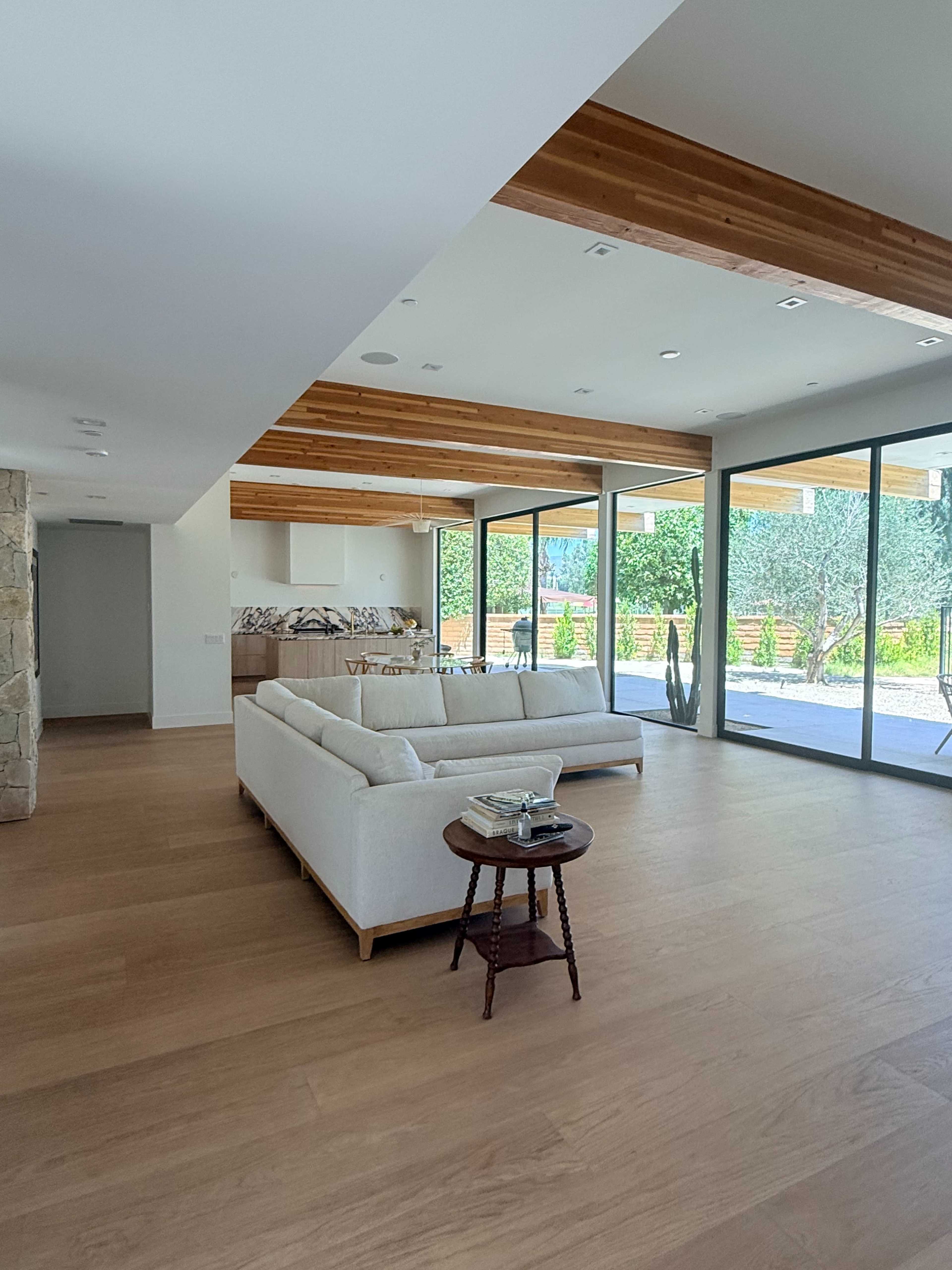 A spacious, modern living area features a light-colored sectional sofa, wooden ceiling beams, and large glass sliding doors that open to an outdoor space.