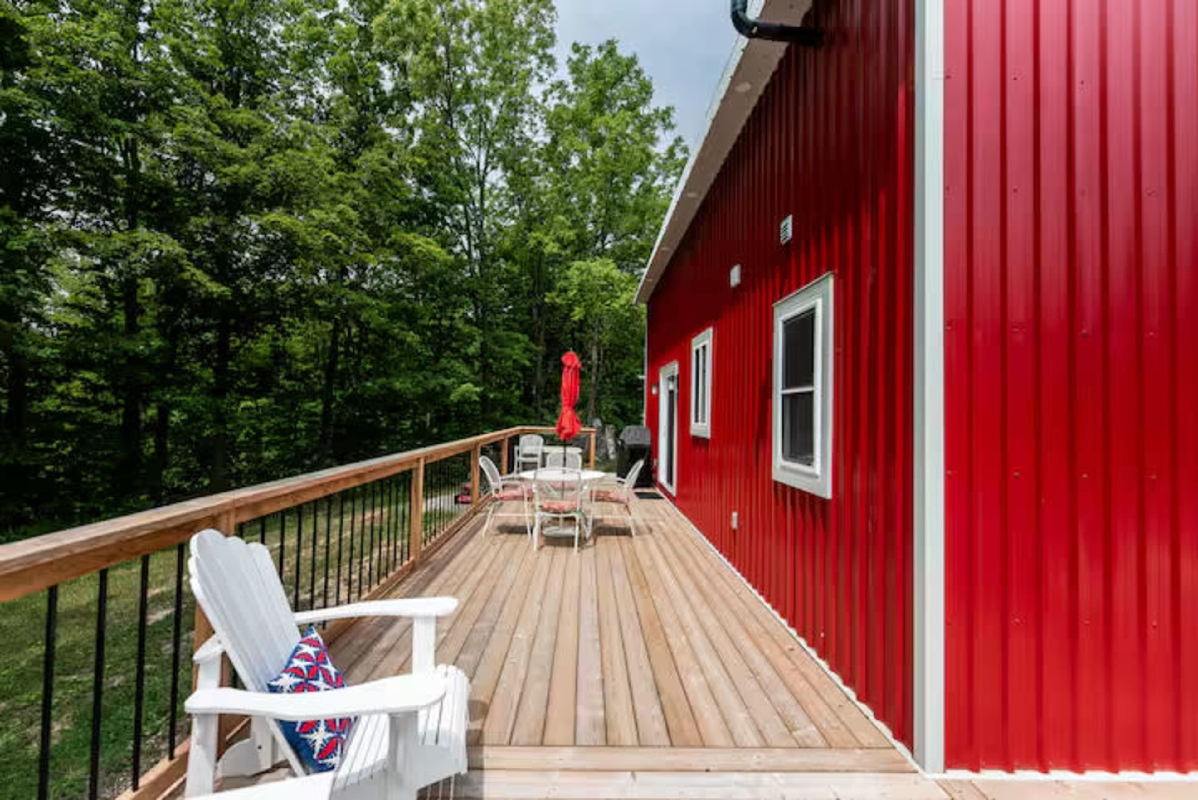 A red metal building features a wooden deck with white outdoor furniture and a view of surrounding trees.