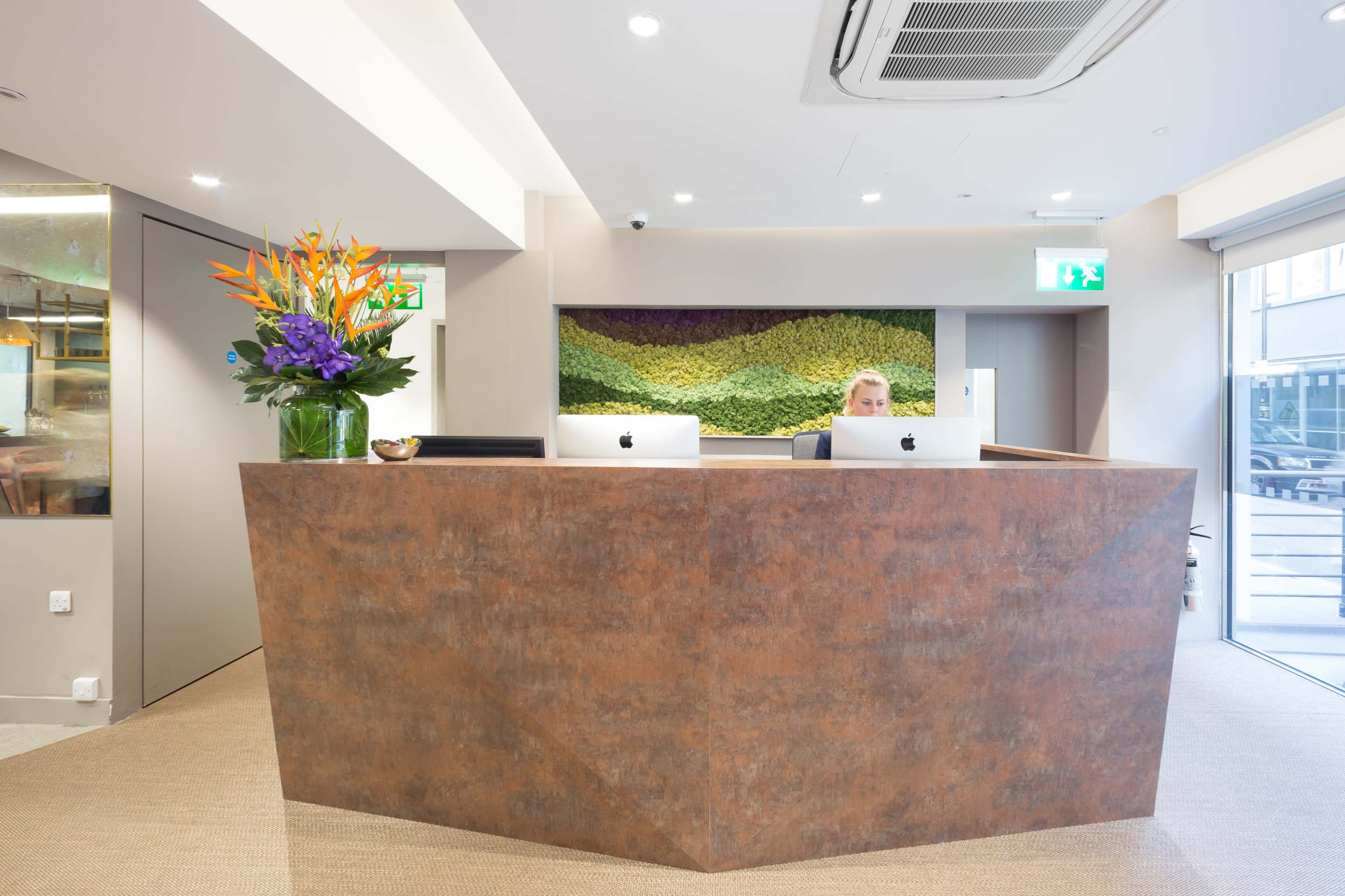 A modern reception area features a geometric front desk with two Apple computers and a flower arrangement, complemented by a green wall art installation.