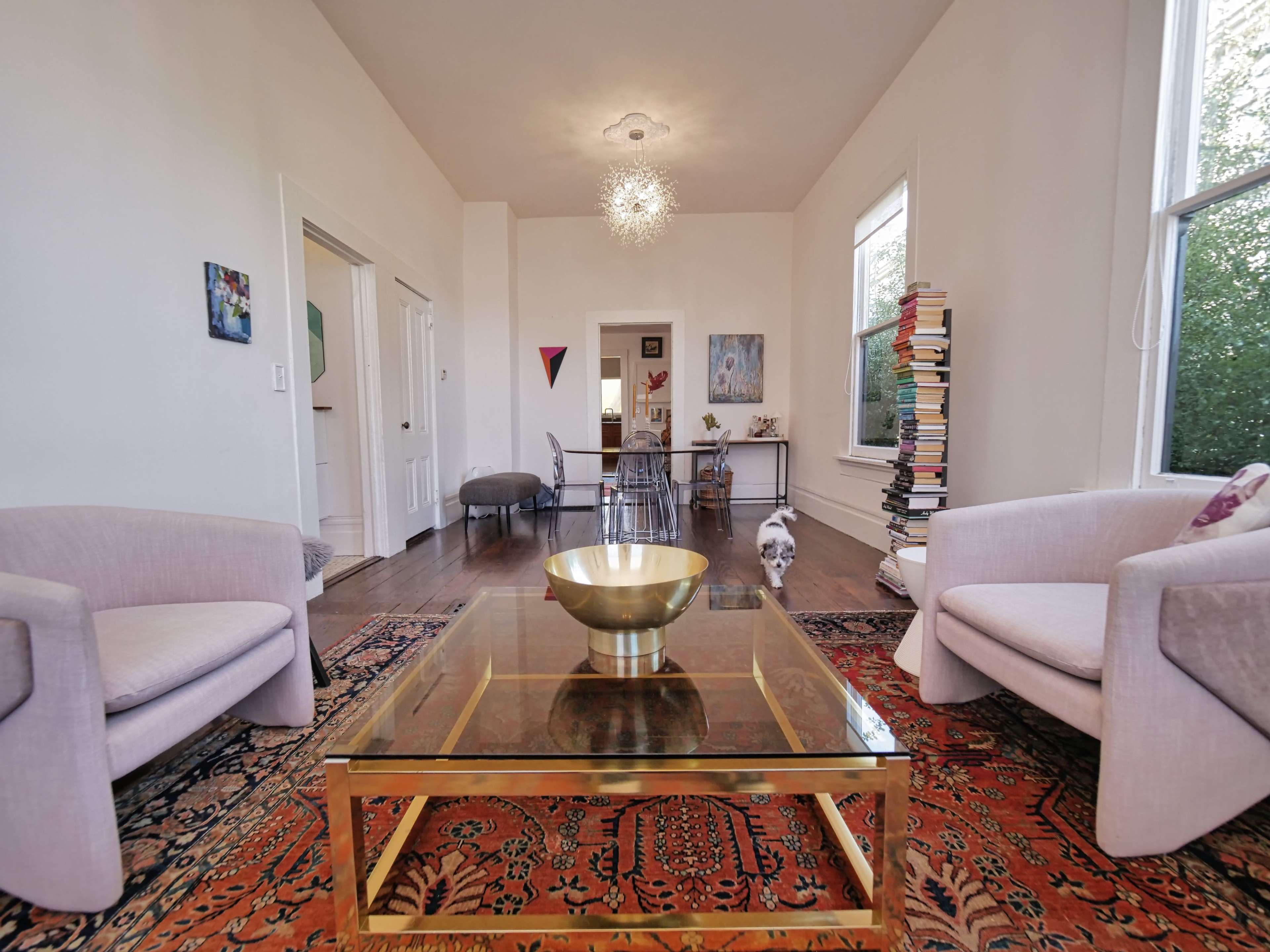 The image shows a living room with two light-colored armchairs, a glass coffee table, and a bookshelf filled with books, all illuminated by a chandelier and natural light from large windows.