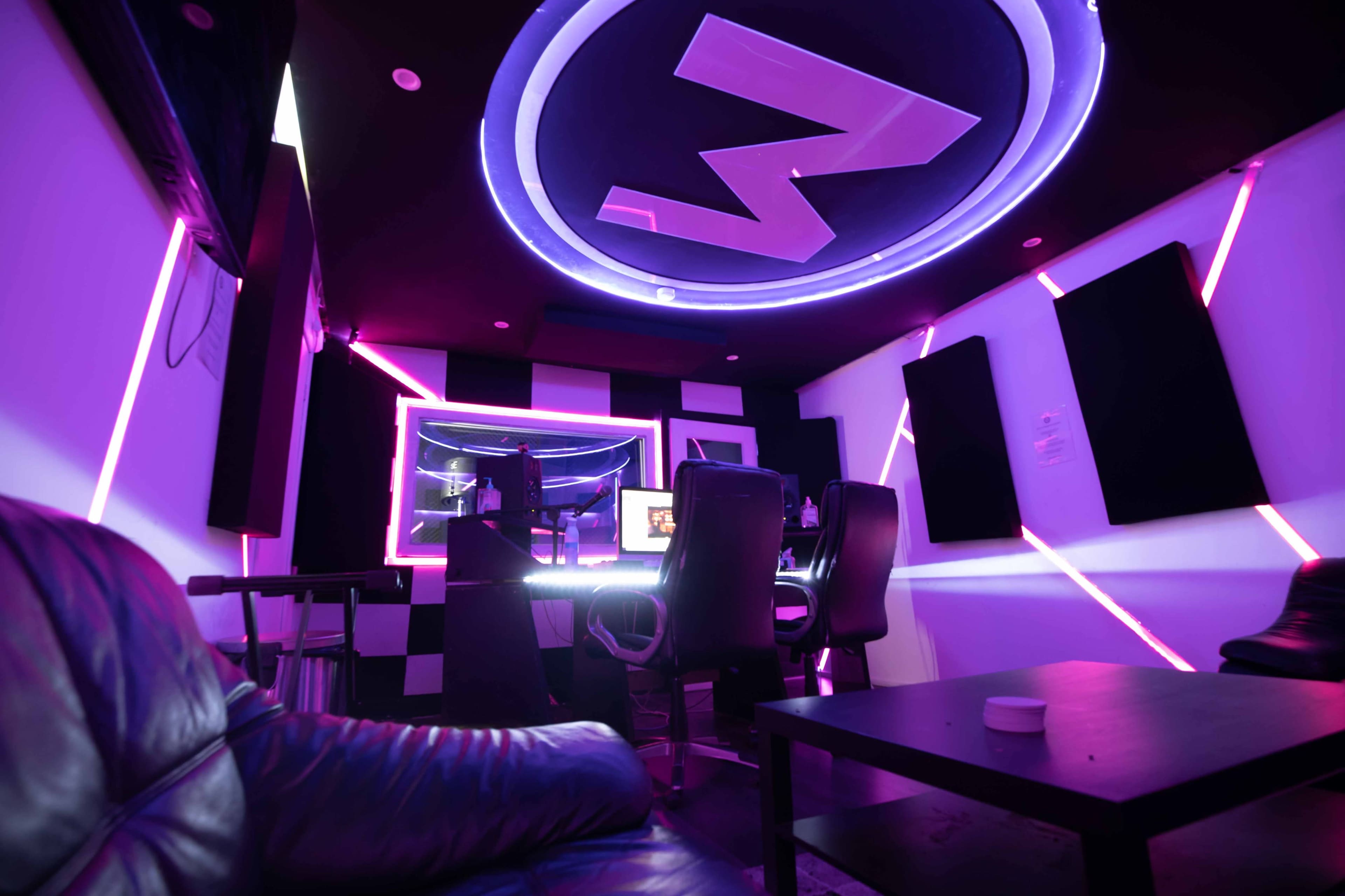 A modern recording studio with purple neon lighting, black and white décor, and two chairs positioned around a central desk.