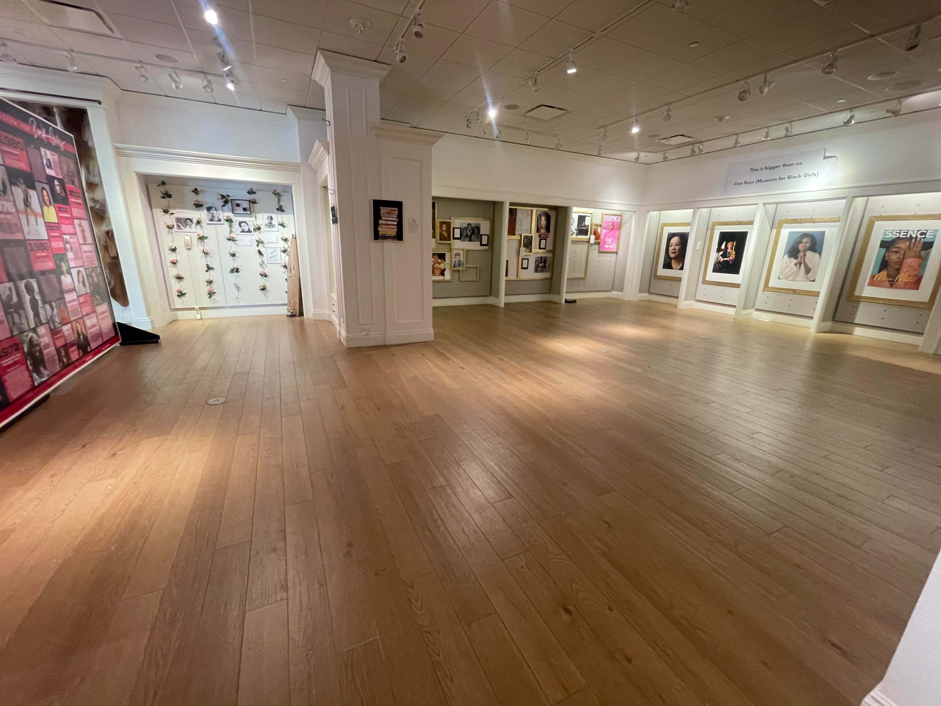 A spacious gallery with wooden flooring and walls displaying various framed artworks and photographs.
