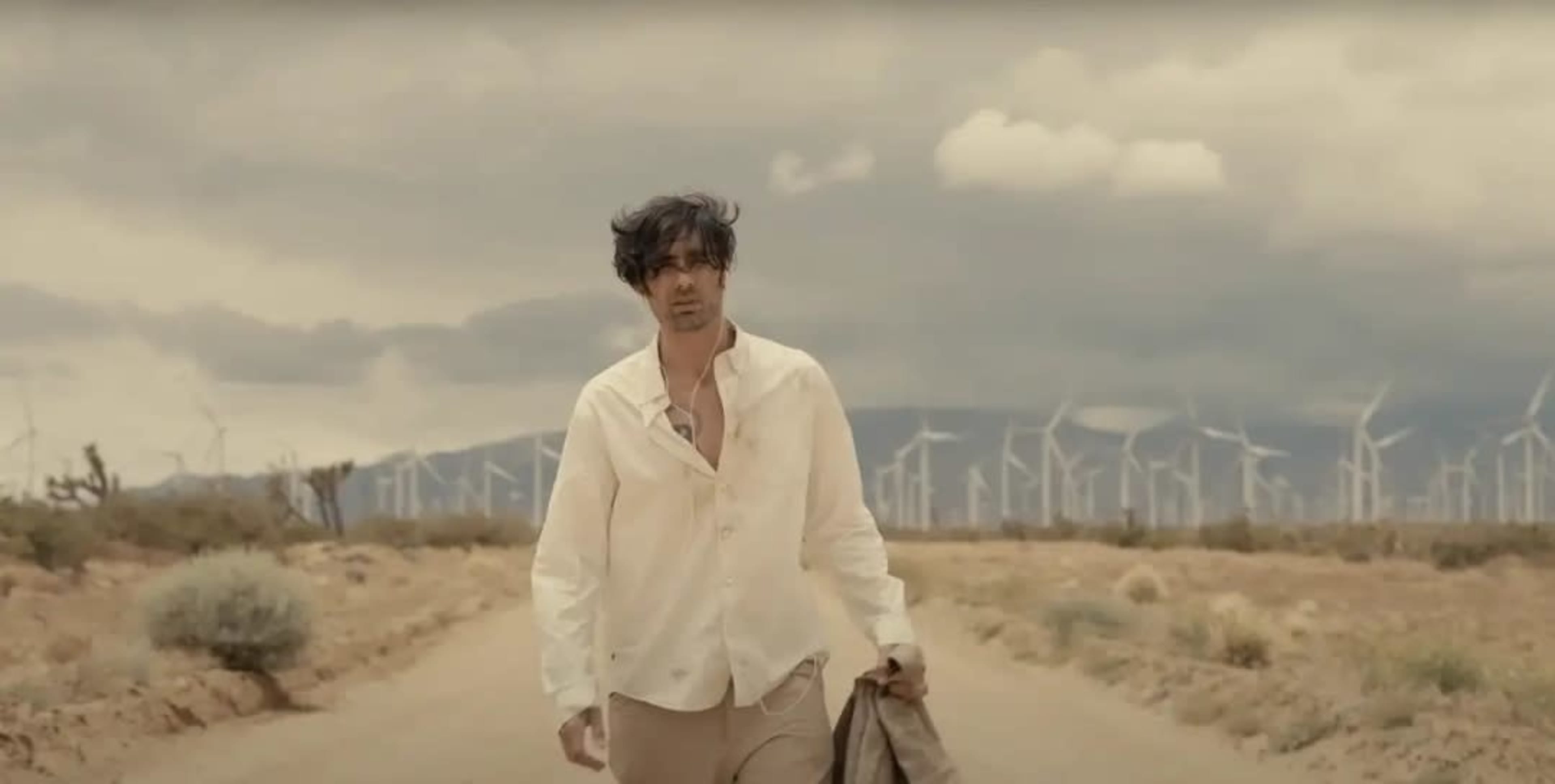 A man walks down a dusty road in a desert landscape, with wind turbines visible in the background under a cloudy sky.
