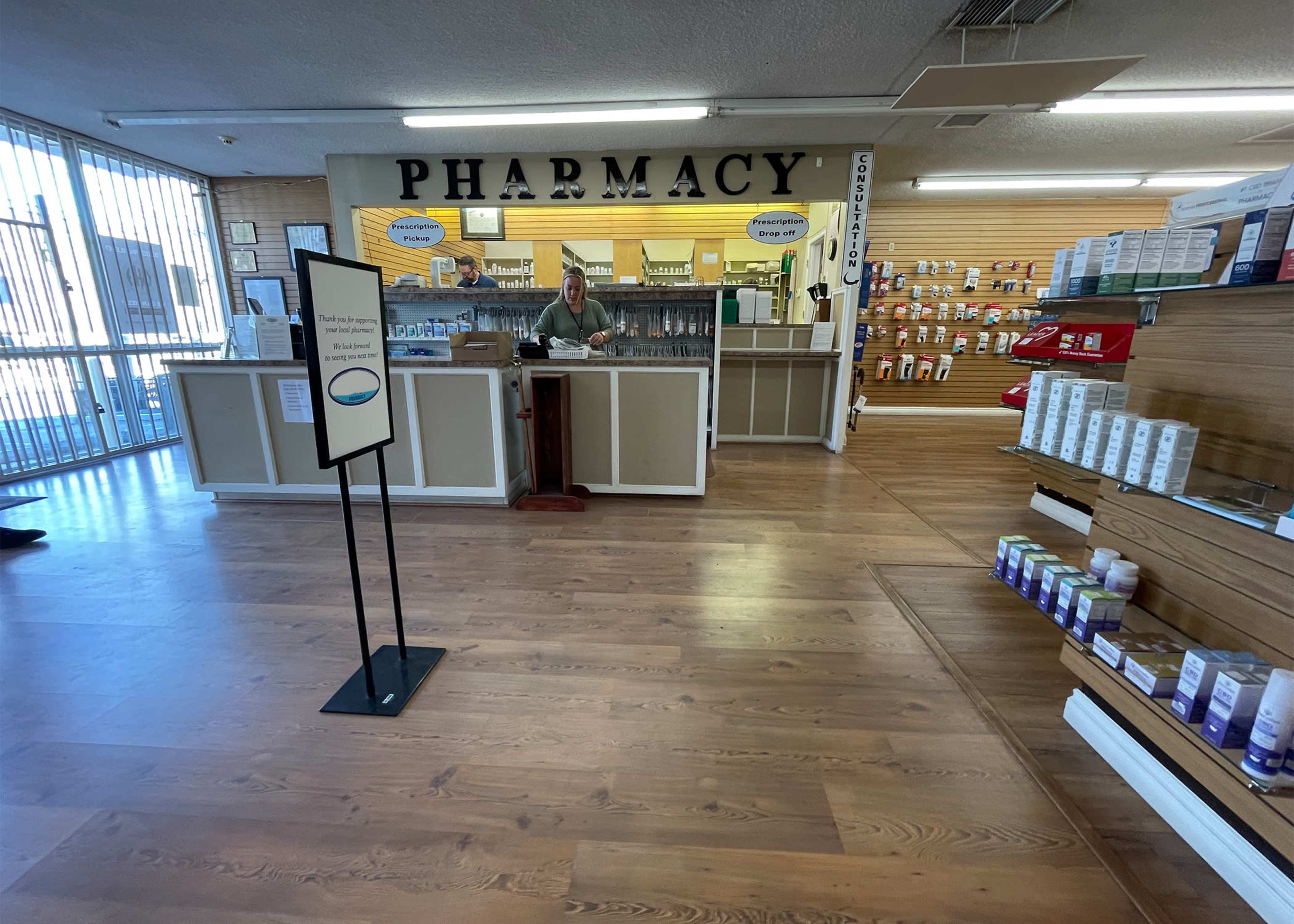 A pharmacy interior features a service counter with a staff member, surrounded by shelves of products and a large sign that reads "PHARMACY."