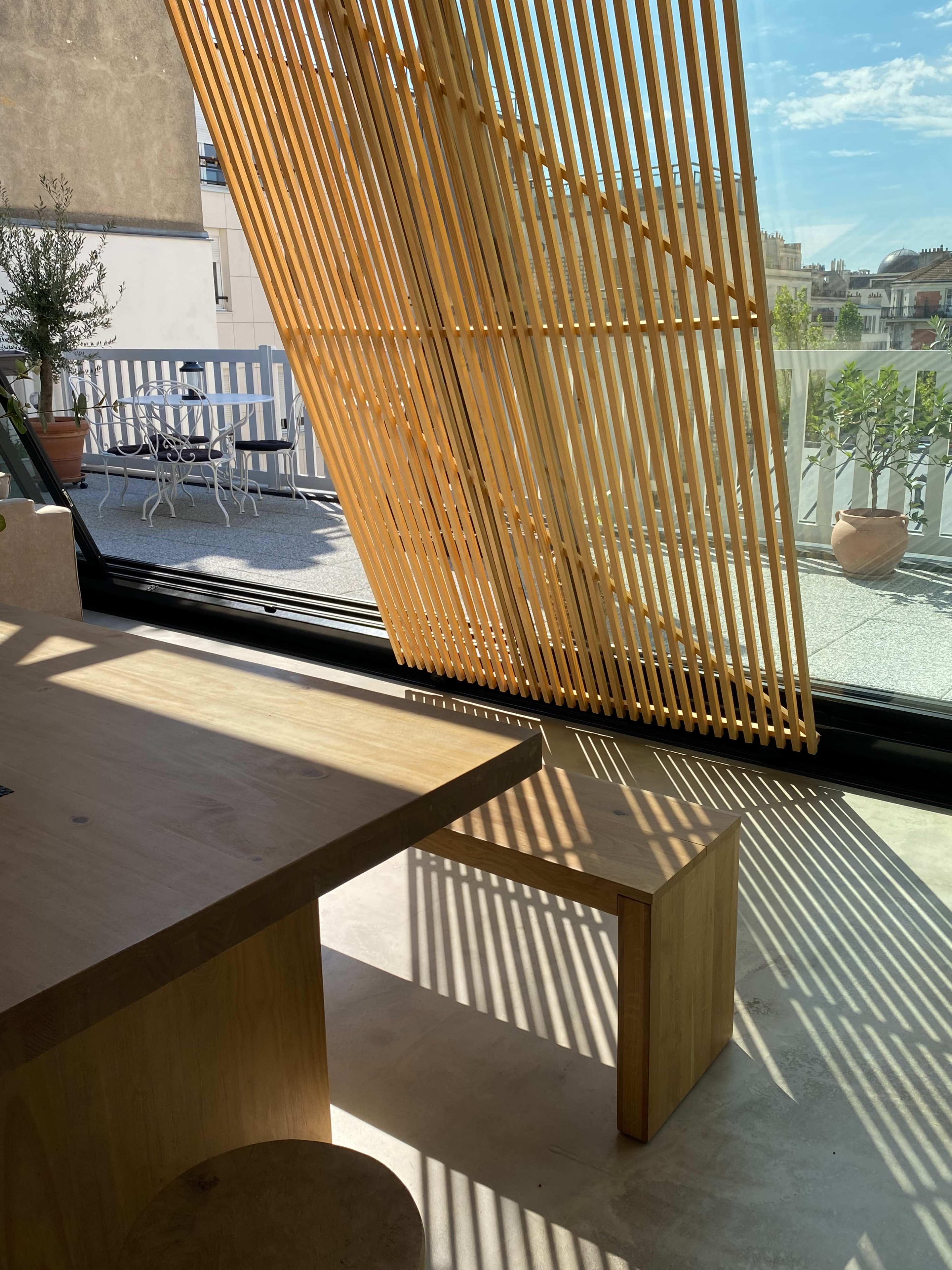 A modern interior space with a wooden table and a bench, illuminated by sunlight filtering through slatted wooden panels, with an outdoor terrace visible in the background.
