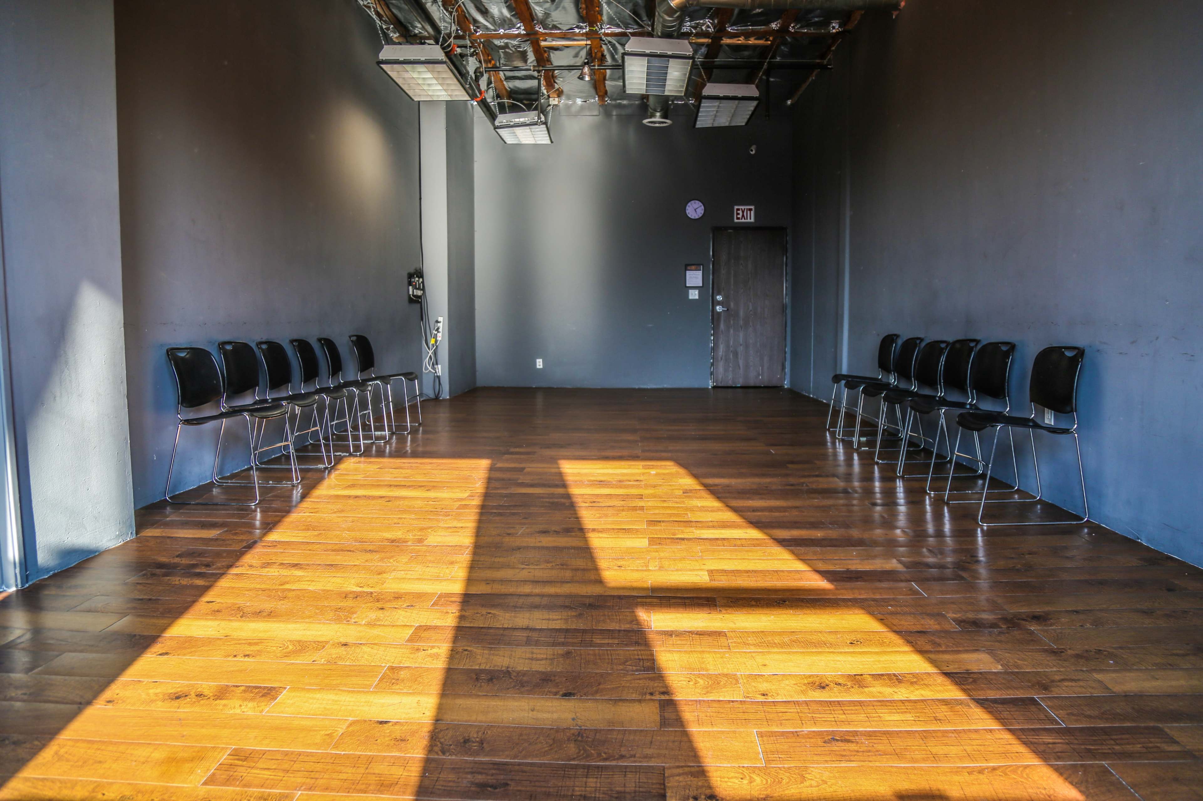 The image shows an empty room with wooden flooring, featuring a row of black chairs along one wall and sunlight casting shadows on the floor.