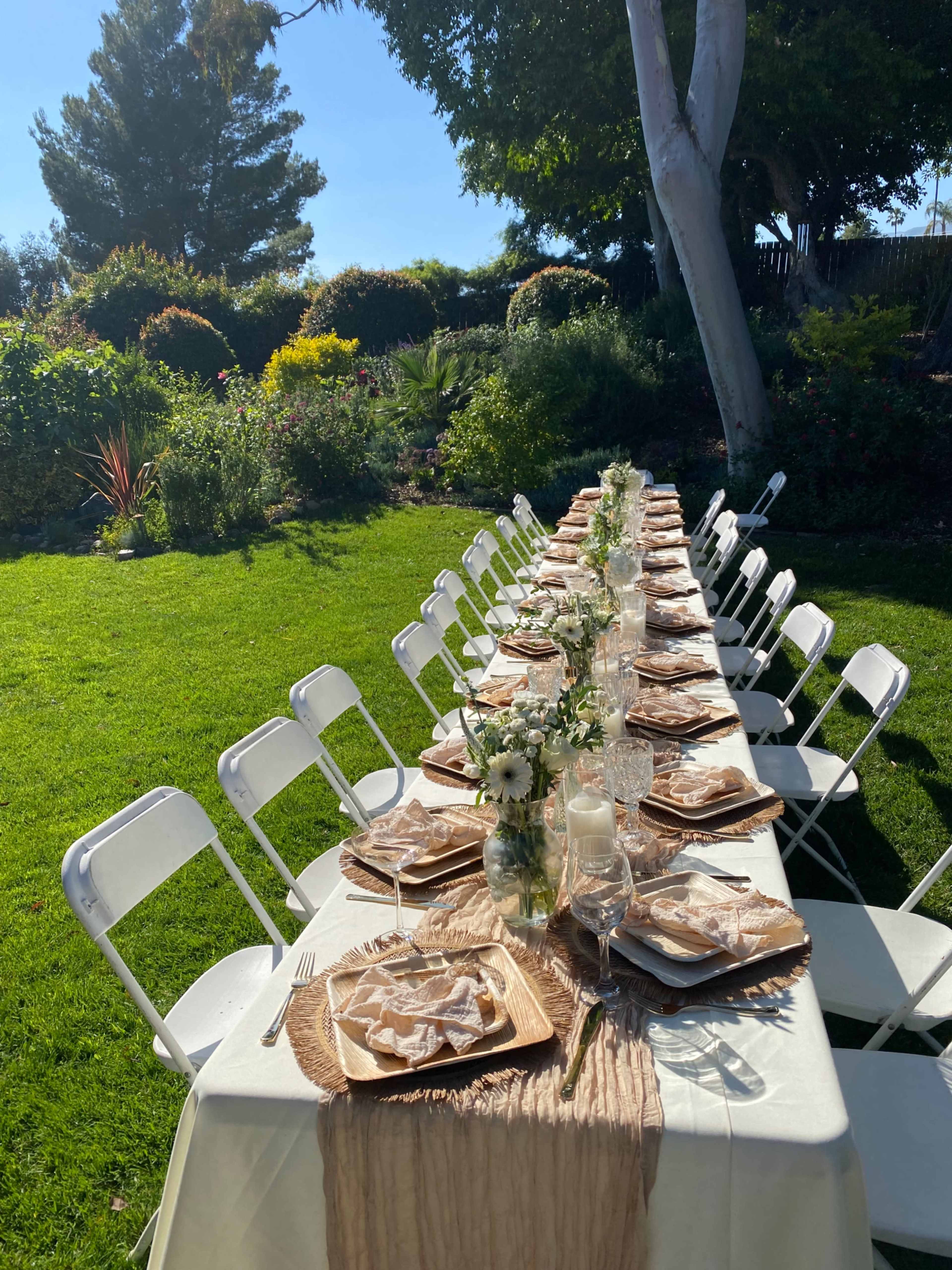 A long, elegantly set table with white chairs is arranged outdoors on a green lawn, surrounded by lush plants and trees.