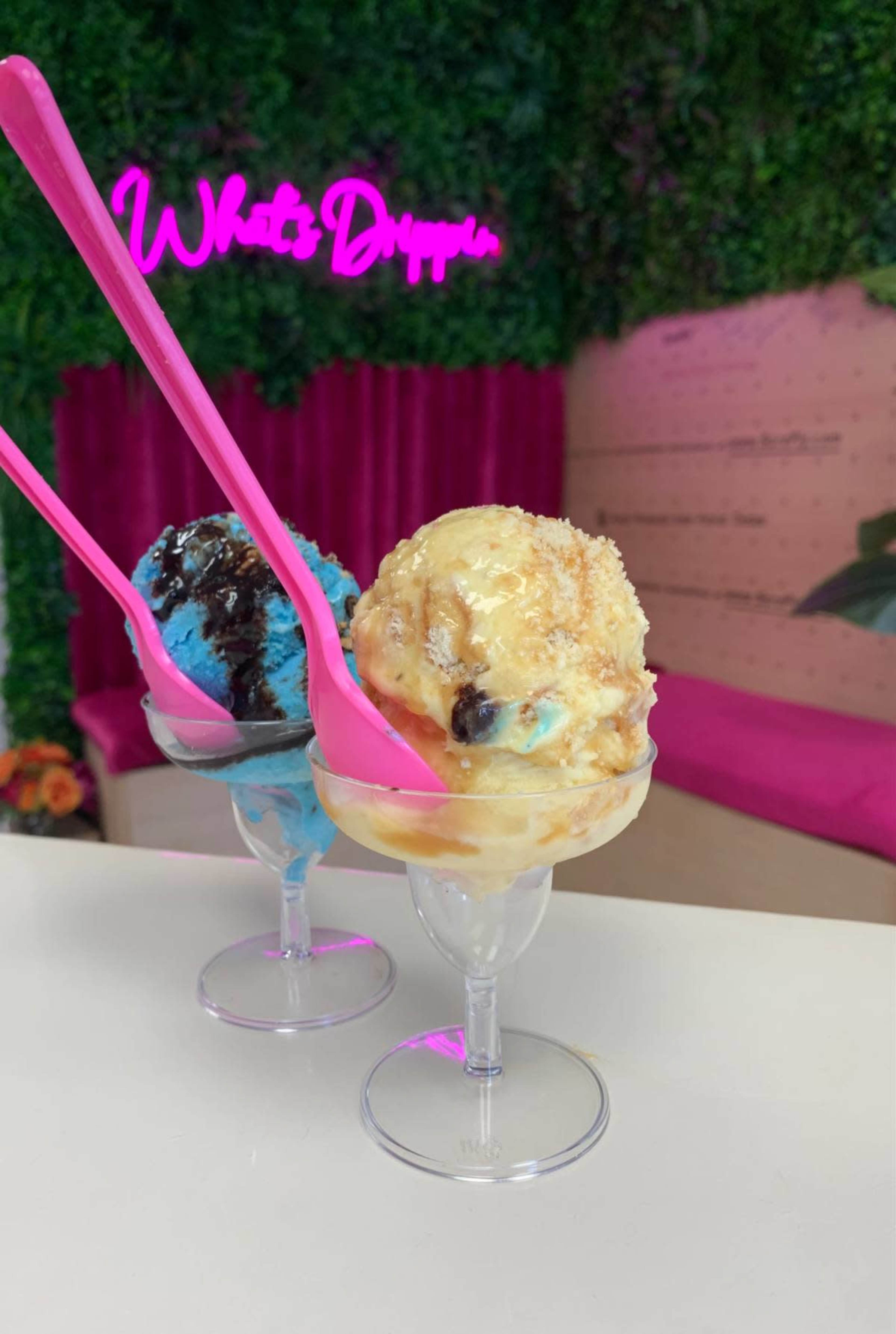 Two ice cream sundaes, one with blue flavor and chocolate syrup and the other with a vanilla base and toppings, are served in clear cups with pink spoons against a backdrop featuring a neon sign.