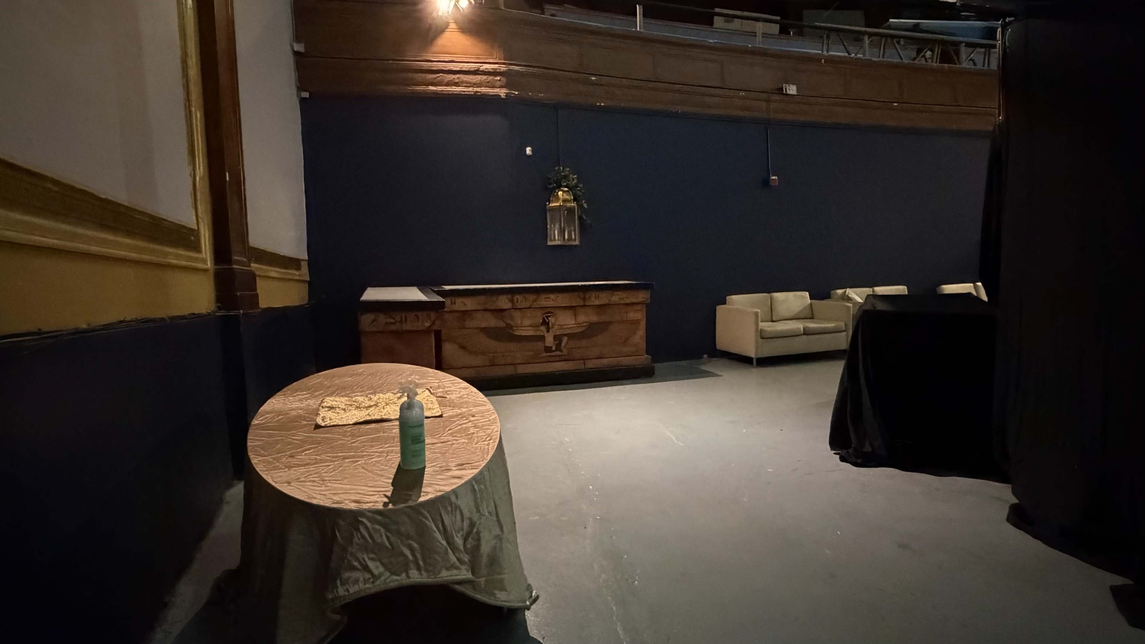 The image shows a sparsely furnished room with a wooden chest, a table covered with a cloth, and a sofa against a dark blue wall.