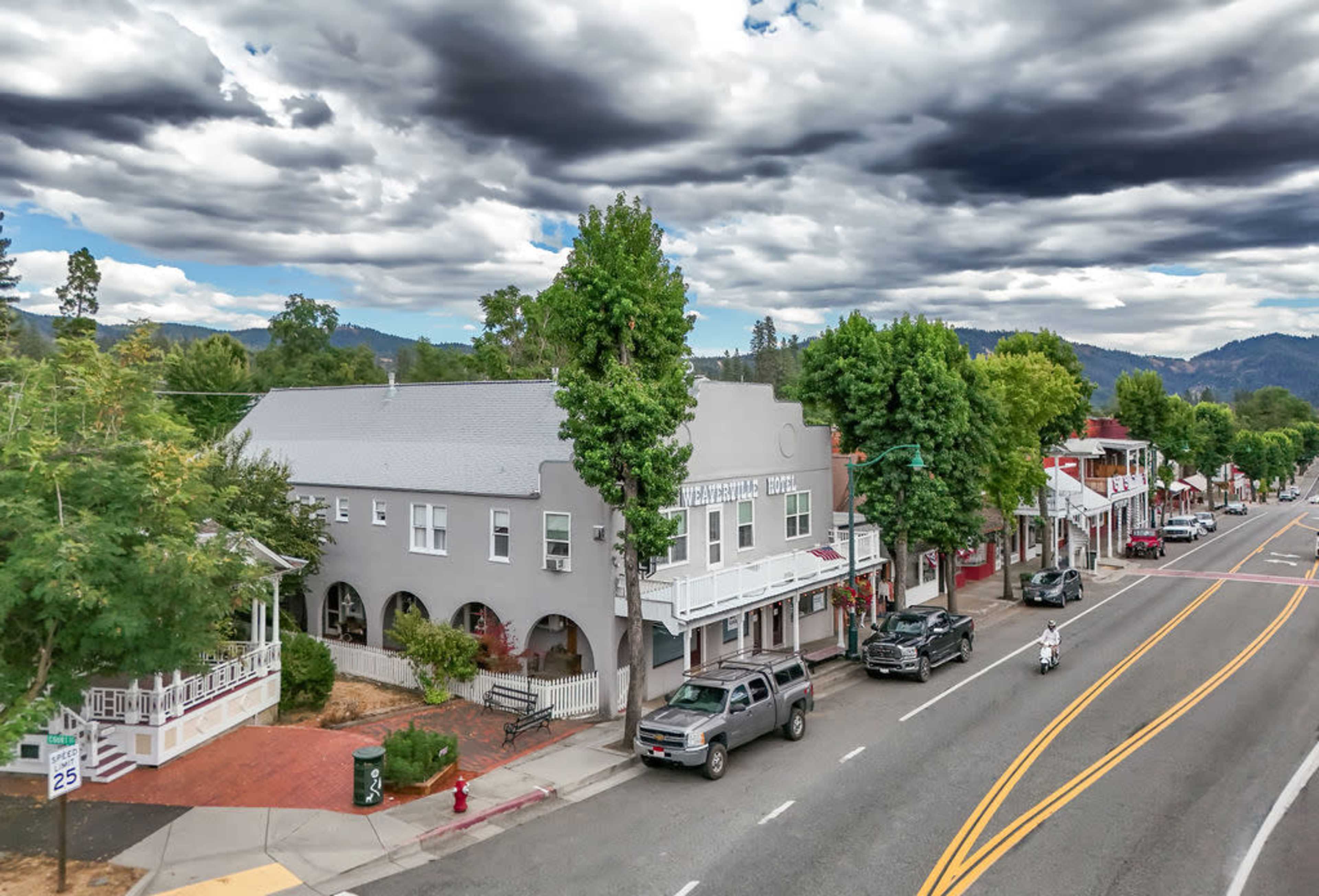 A street scene features a row of buildings alongside a two-lane road, with trees lining the area and vehicles parked near the curb.
