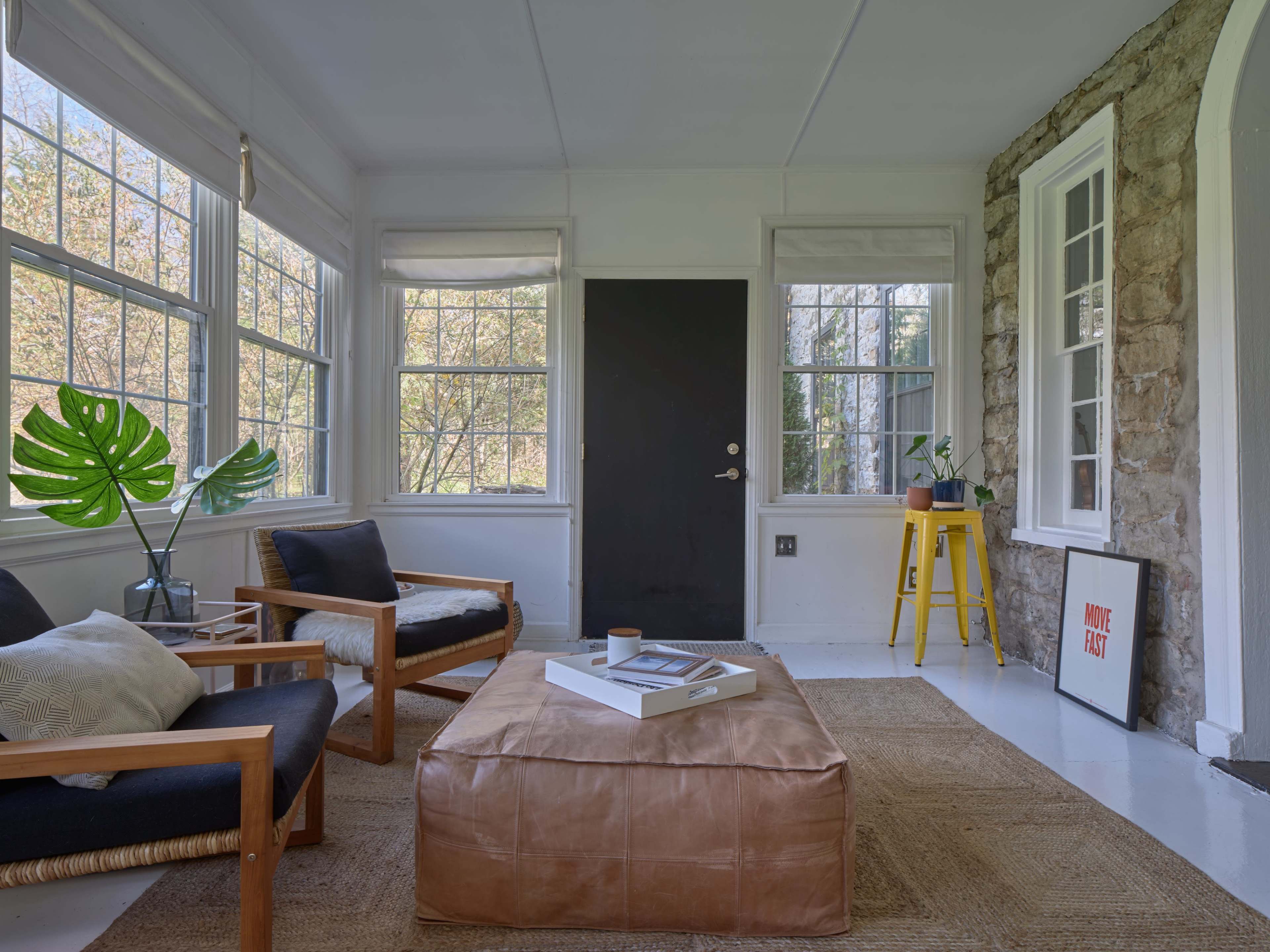 A sunlit room with large windows, featuring two wooden armchairs, a coffee table on a woven rug, and a door leading outside.