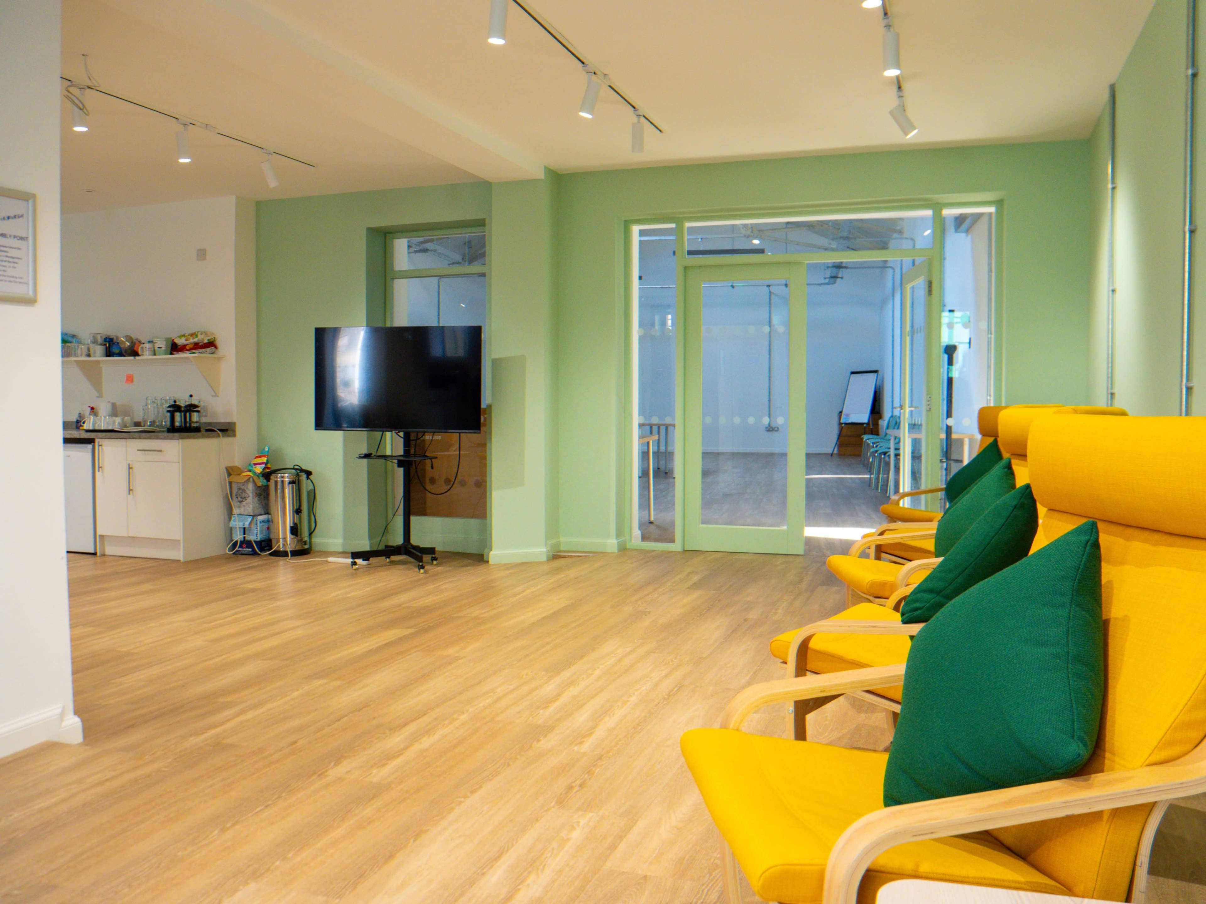 The image shows a brightly lit, modern workspace with yellow armchairs, green accent walls, a television, and an open area leading to another room.