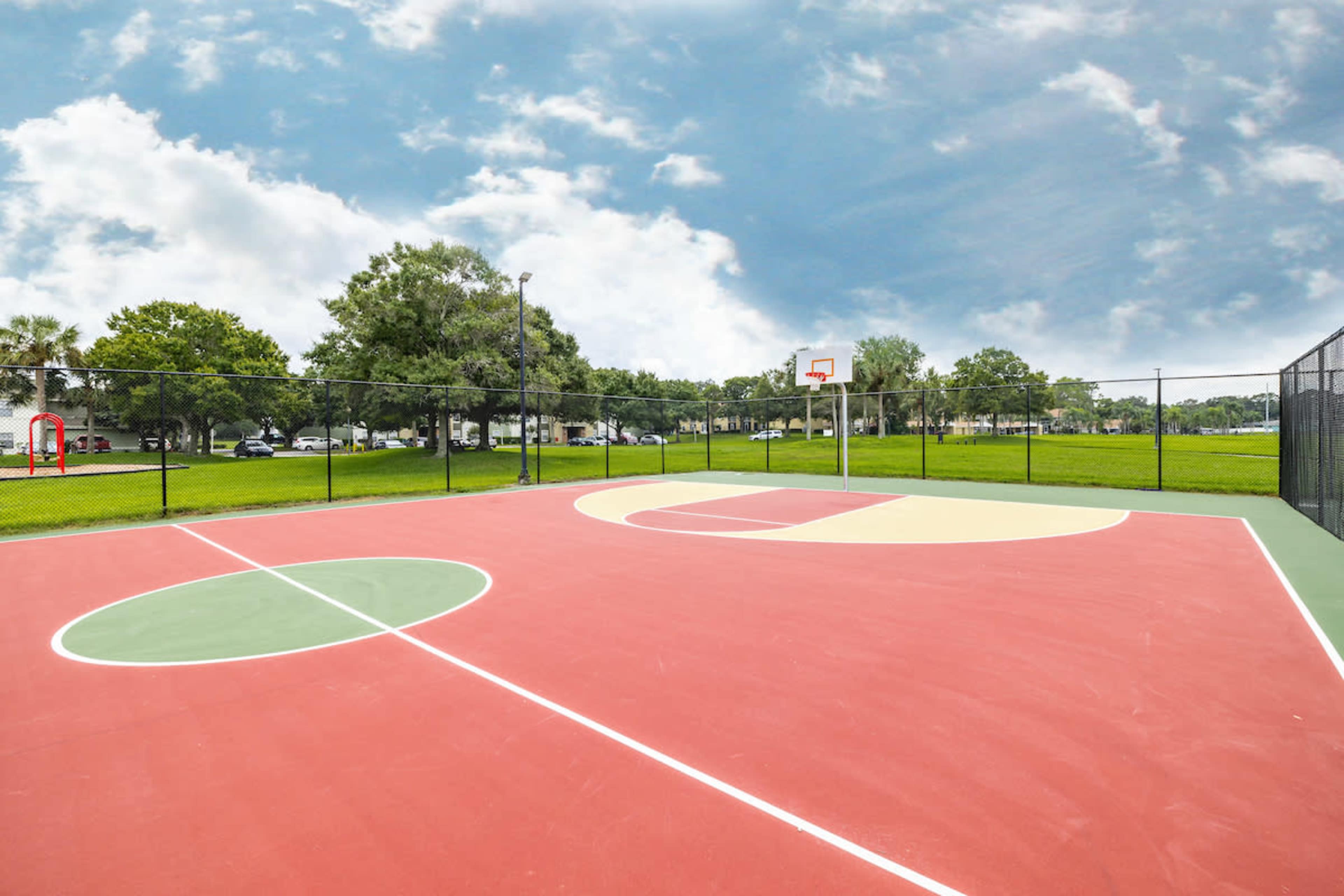 A vacant outdoor basketball court surrounded by grass and trees, with a clear sky overhead.