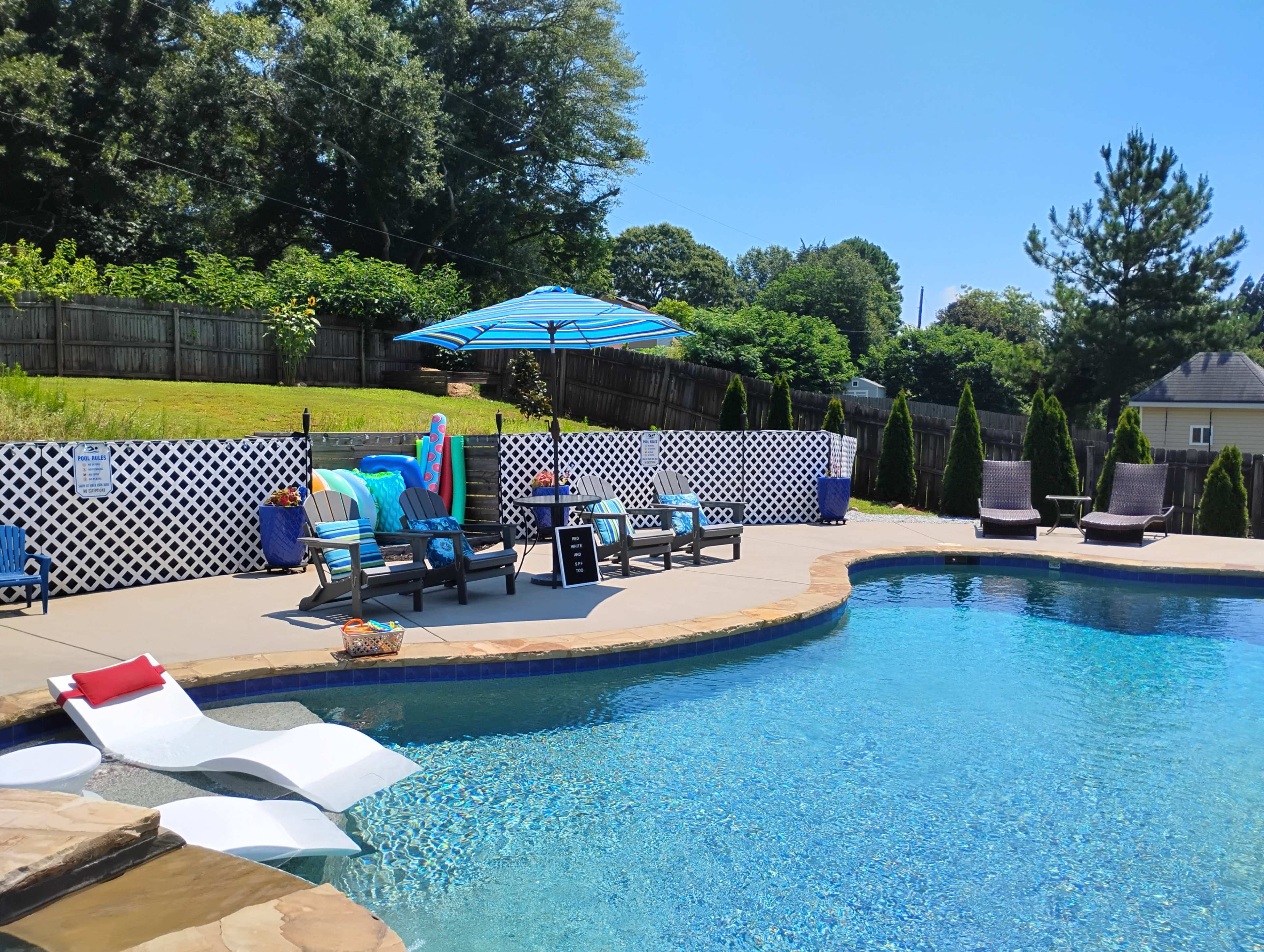 A swimming pool area features lounge chairs and an umbrella against a backdrop of greenery and a wooden fence.