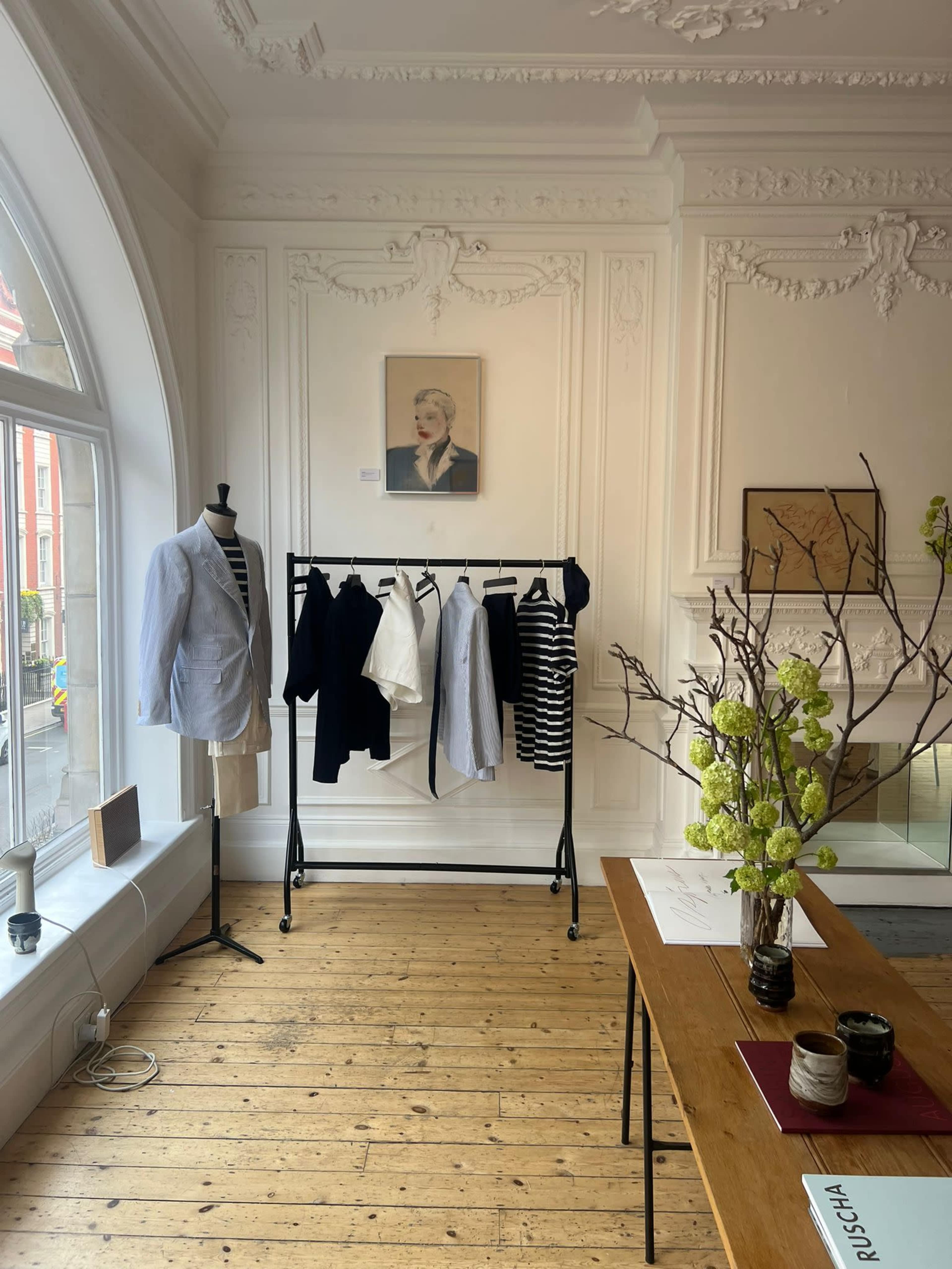 The image shows a boutique interior featuring a mannequin dressed in a light blue shirt, a clothing rack with various garments, and a decorative table with a vase of flowers.