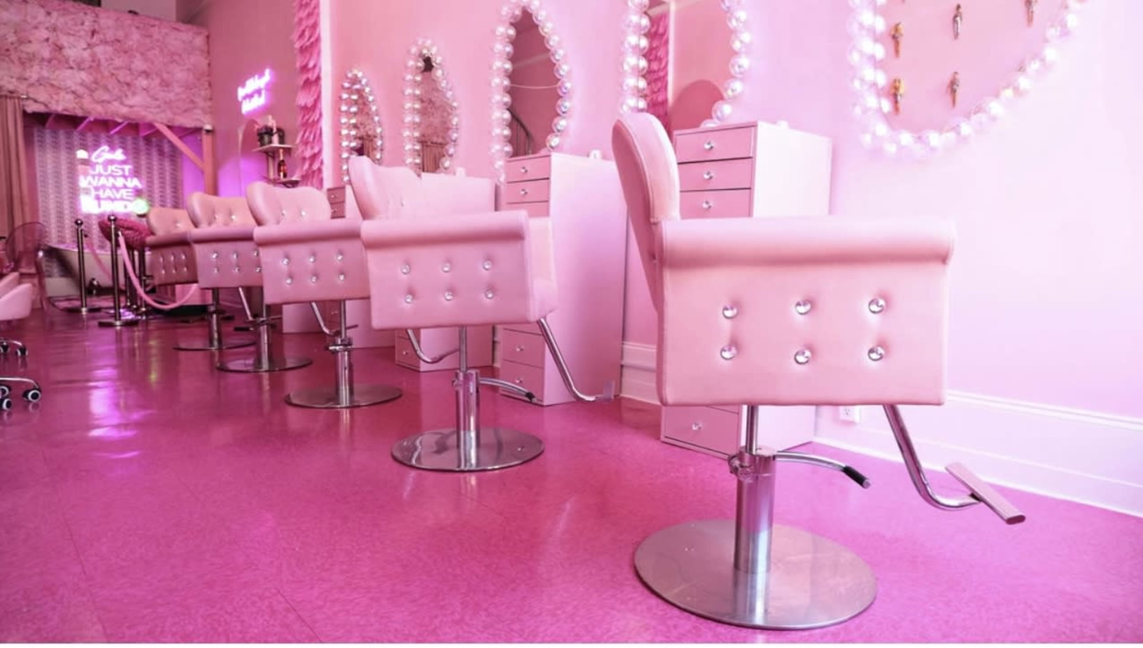 A row of pink salon chairs positioned in front of mirrored stations in a brightly lit salon with a pink decor theme.