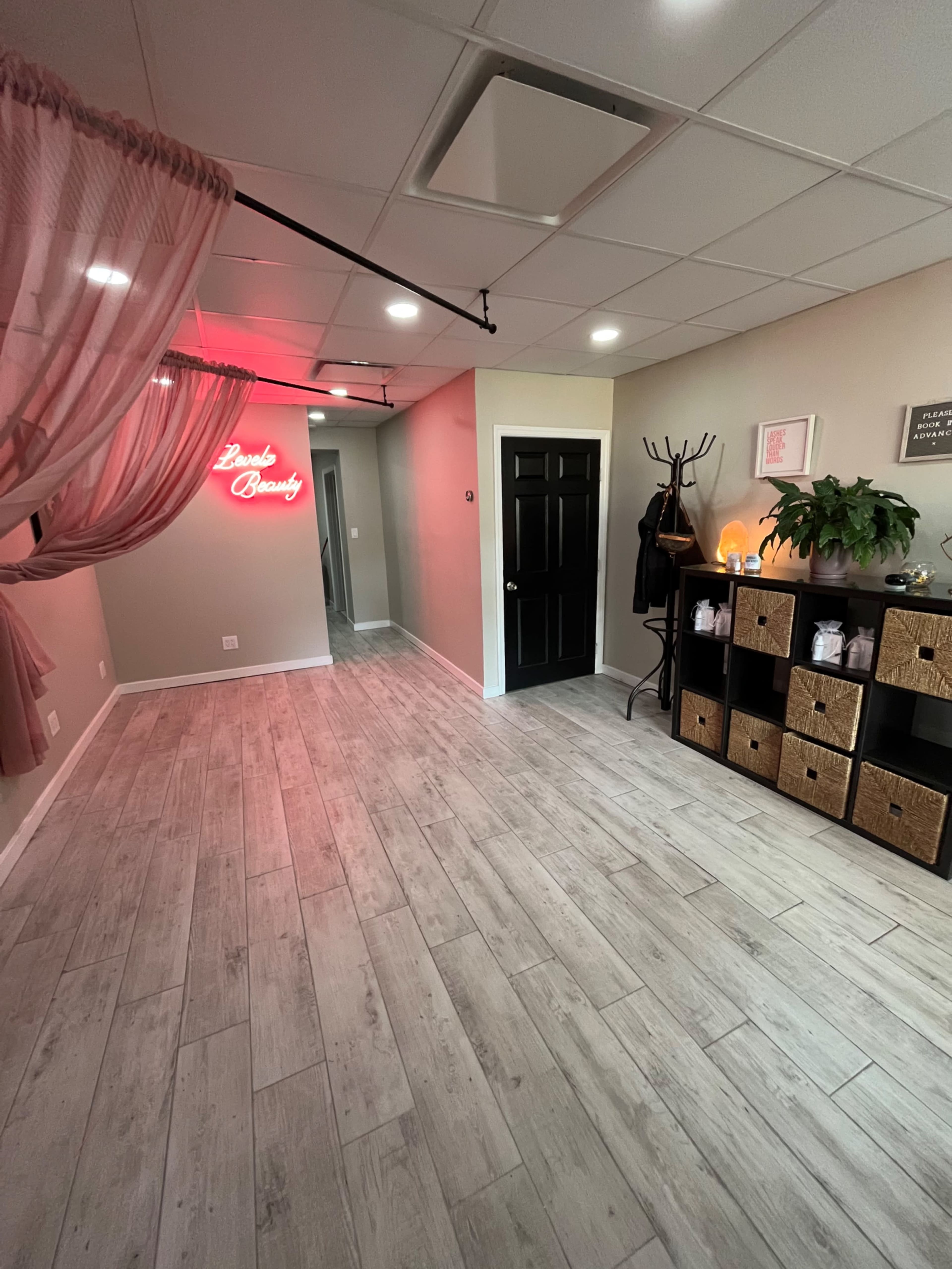 A spacious room with light-colored wooden flooring, a black door, decorative storage boxes, a plant, and a neon sign that reads "Luxe Beauty."