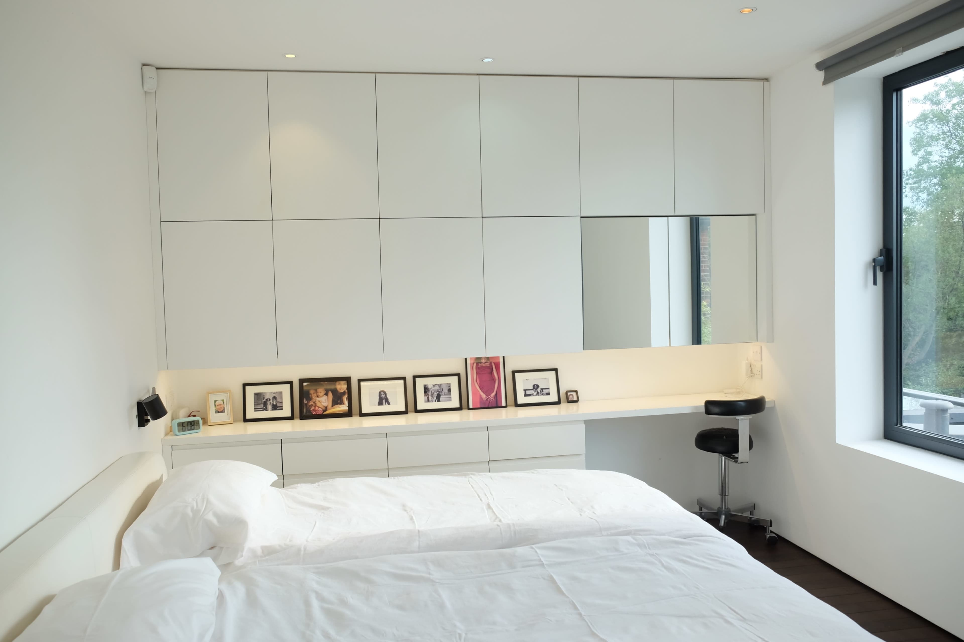 A minimalistic bedroom features a large bed, a row of framed photographs on a white shelf, and a sleek mirror above a small black stool.