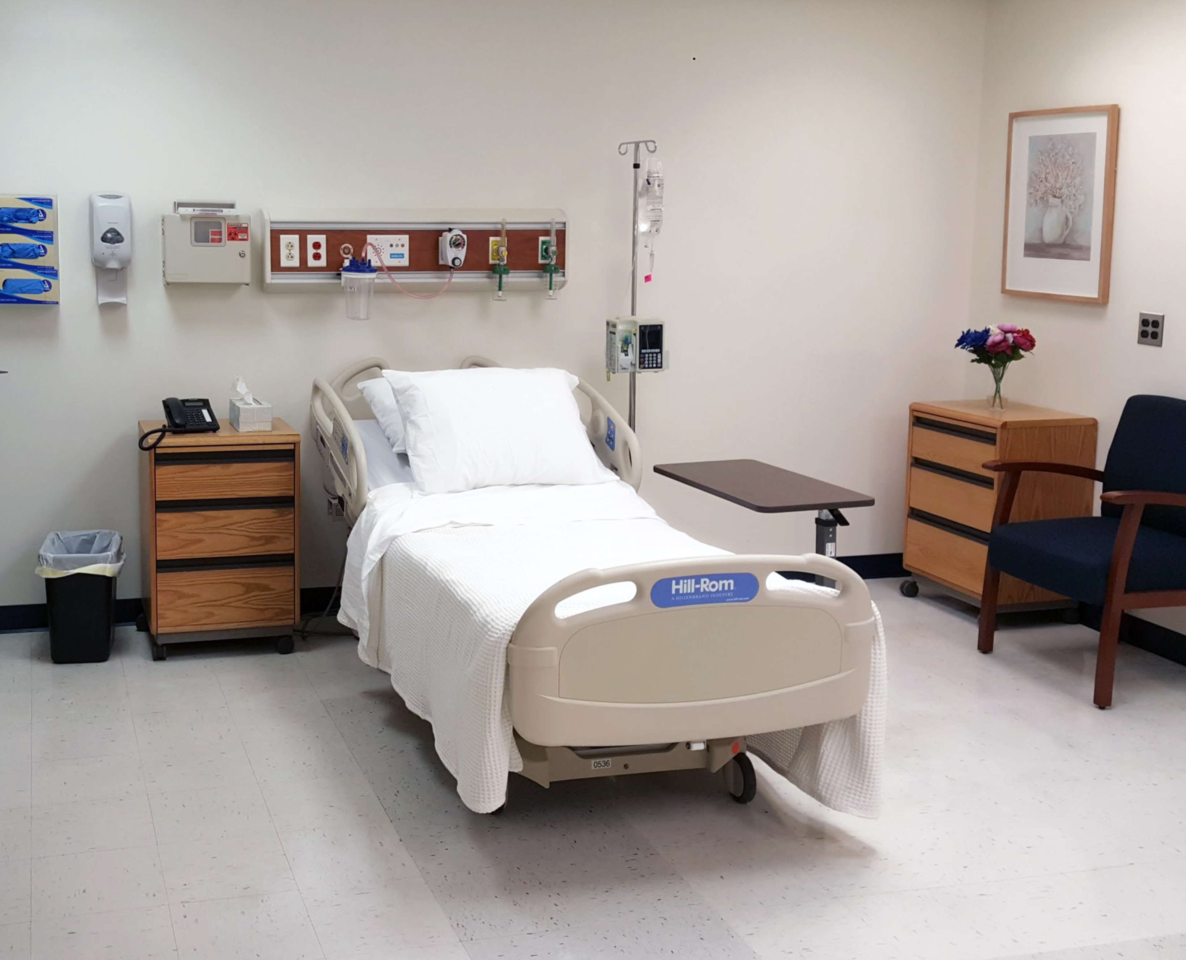 A hospital room featuring a bed with white linens, a bedside table, a chair, and a small bouquet of flowers on a dresser.
