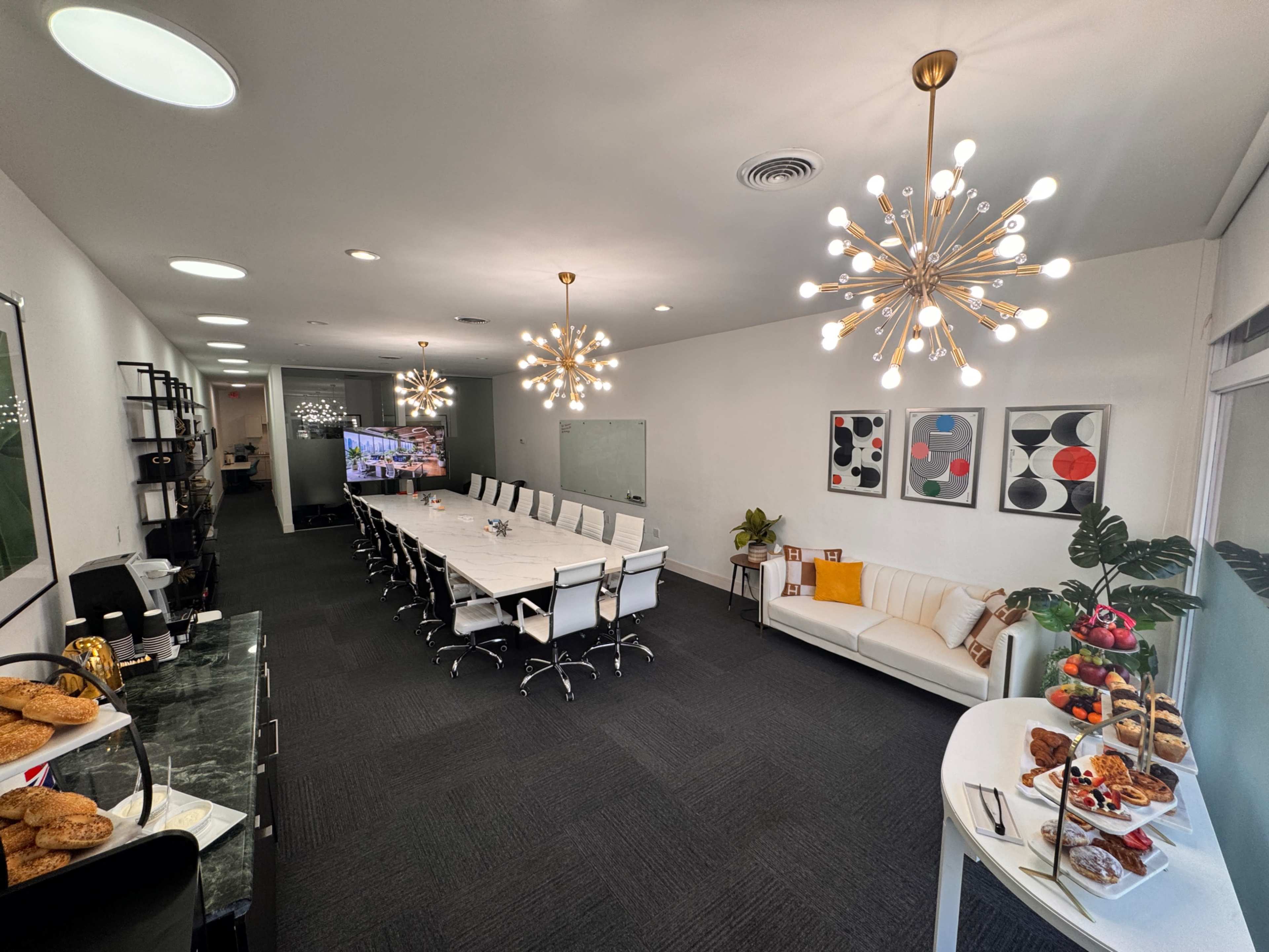 A modern conference room features a long table with chairs, bright lighting, and a snack area displaying various food items.