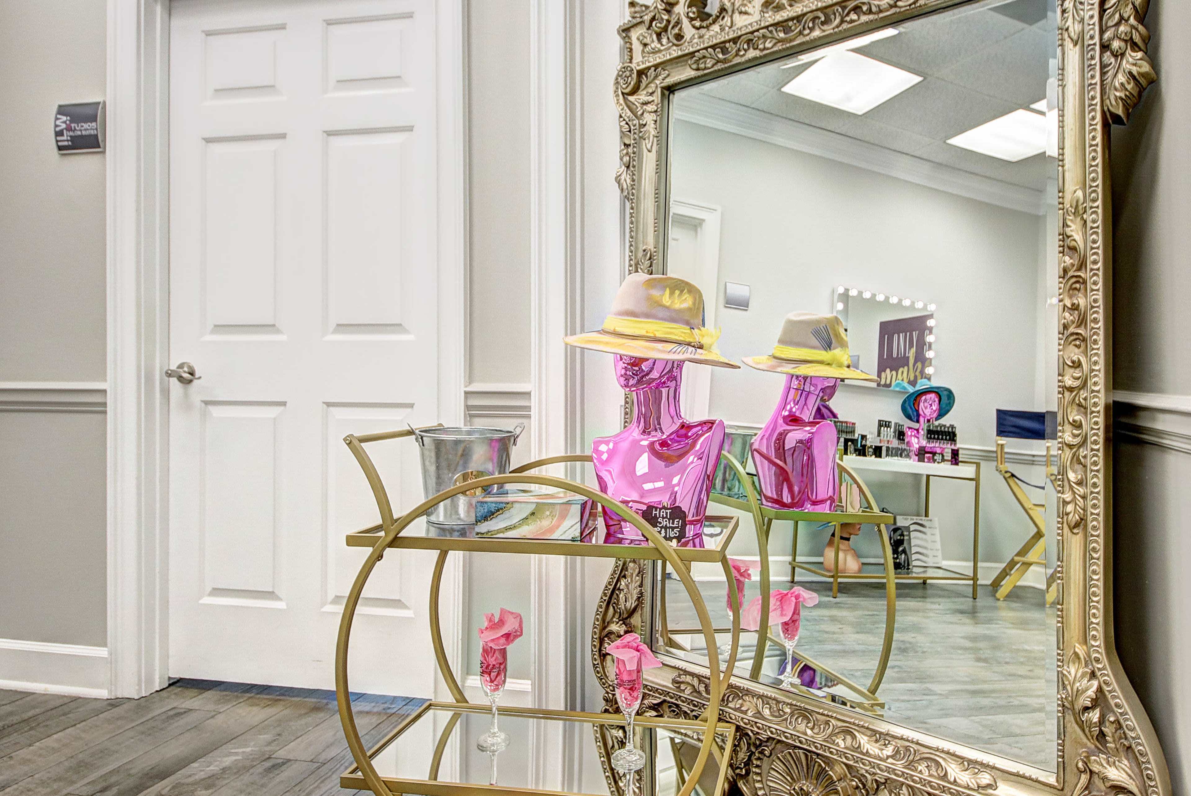 The image showcases a stylish salon interior featuring a large ornate mirror, a pink mannequin with a hat, and a gold rolling cart with various beauty products.