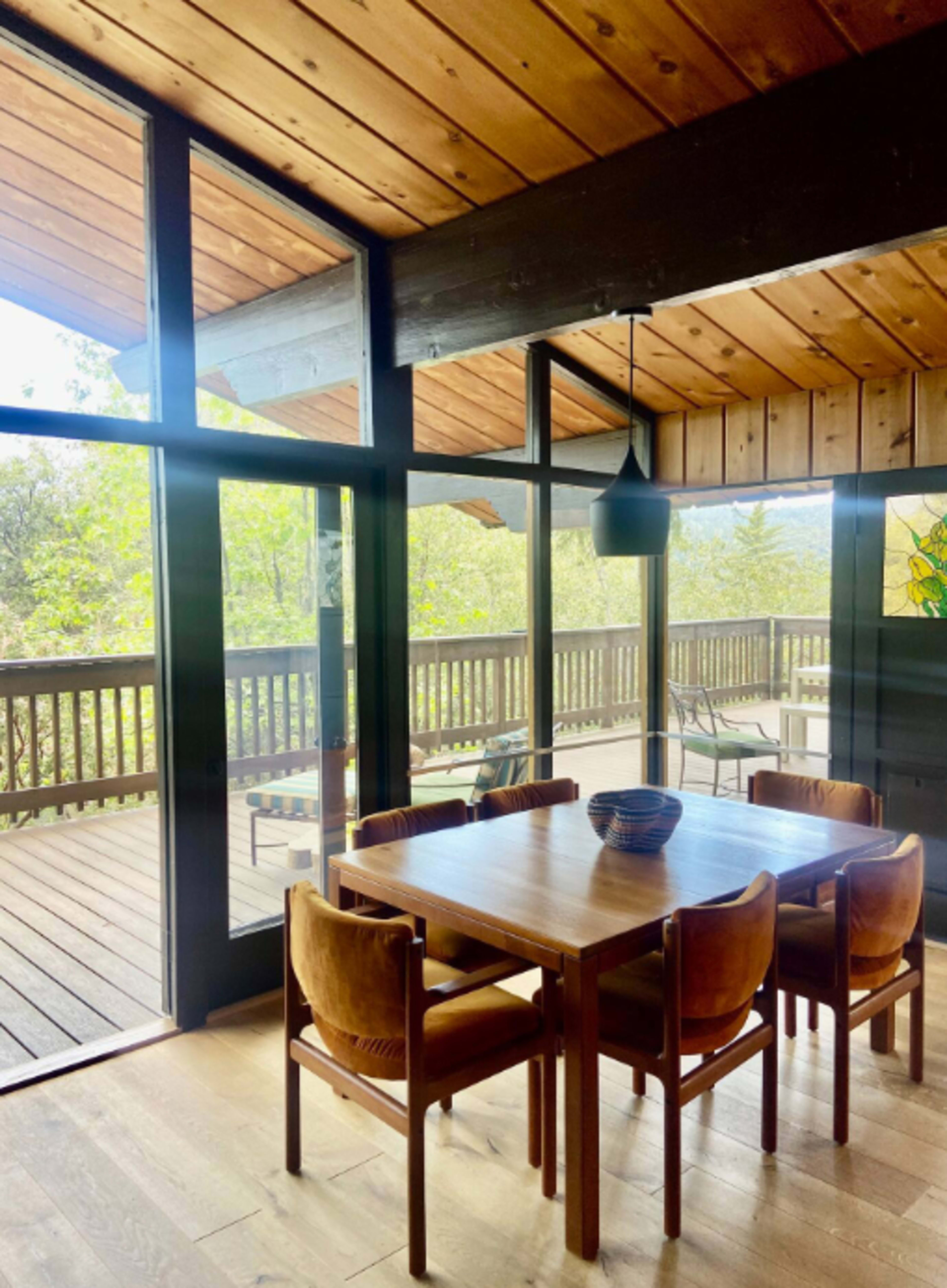 A wooden dining table with four chairs is positioned near large windows that open to a deck surrounded by trees.