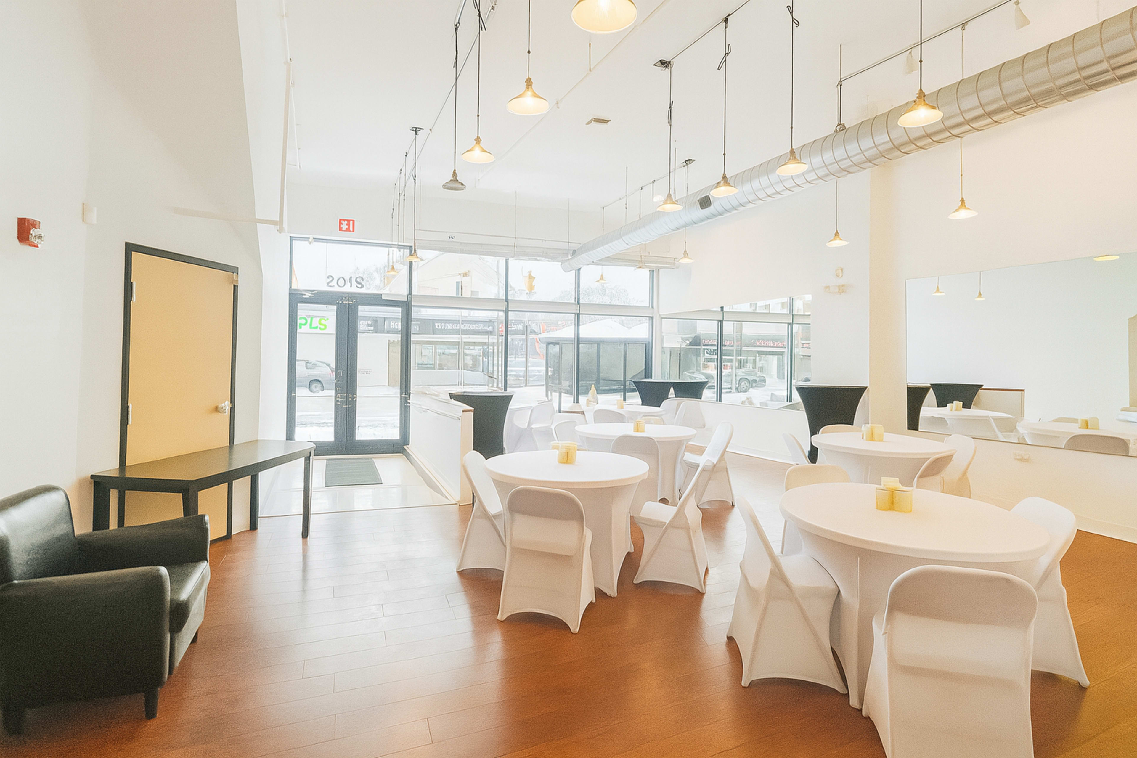 The image shows a spacious, well-lit interior featuring round tables with white tablecloths, modern chairs, and large windows providing a view of the street outside.