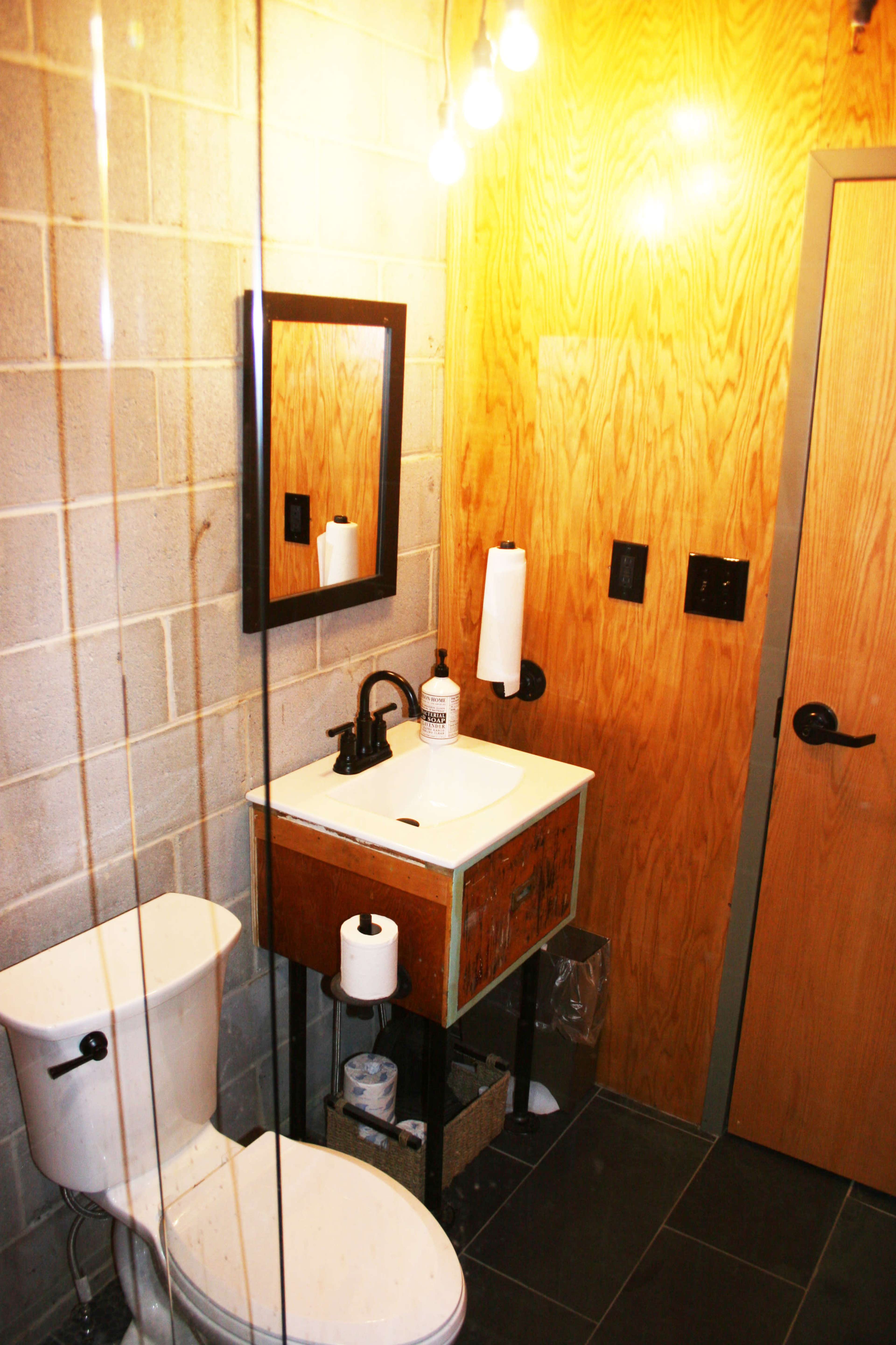 A small bathroom with a toilet, a sink with a wooden cabinet, and a mirror, featuring exposed light bulbs and a wooden wall.
