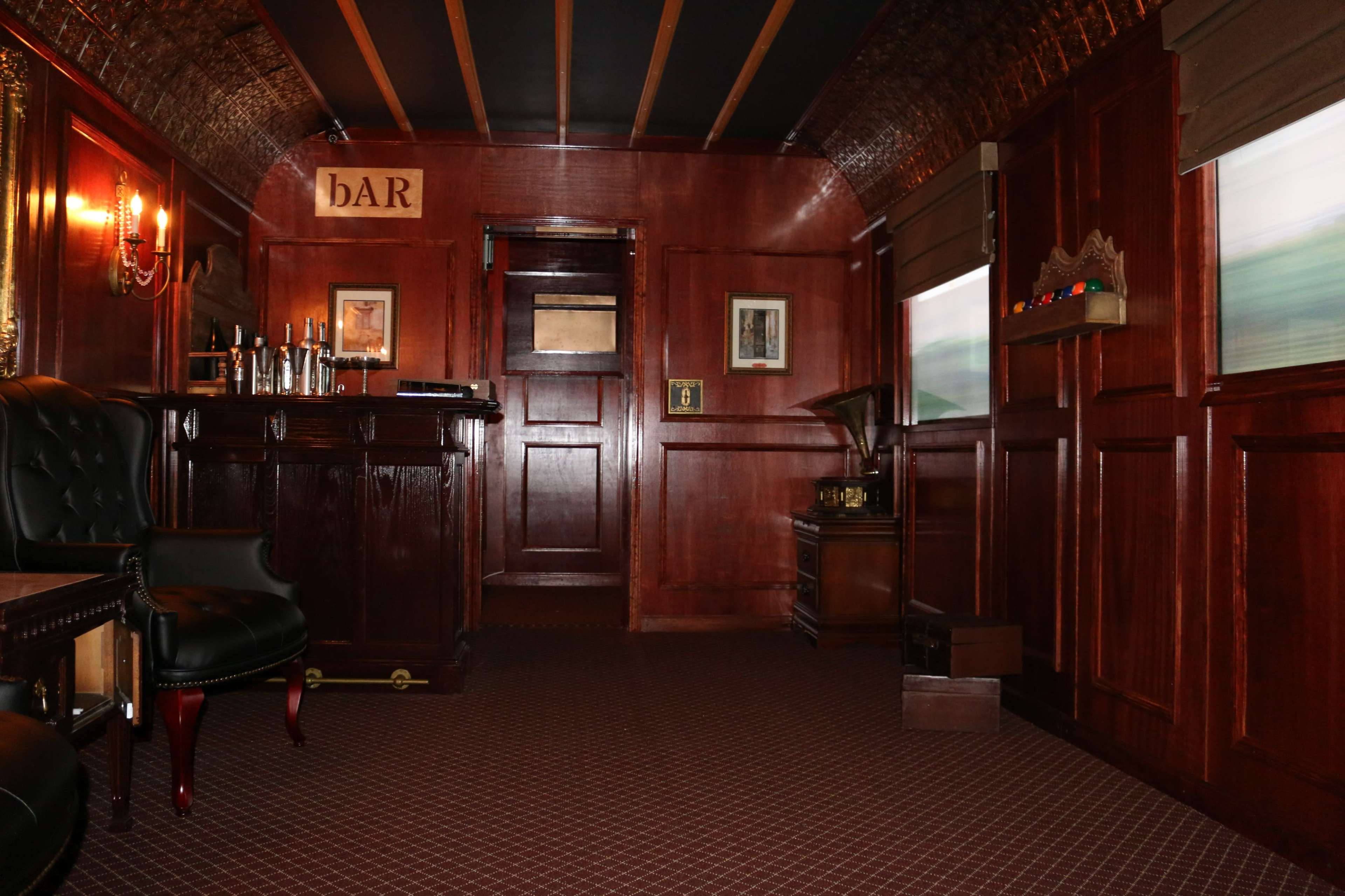 The interior of a vintage train car features dark wood paneling, a bar counter, and a seating area with black leather chairs.