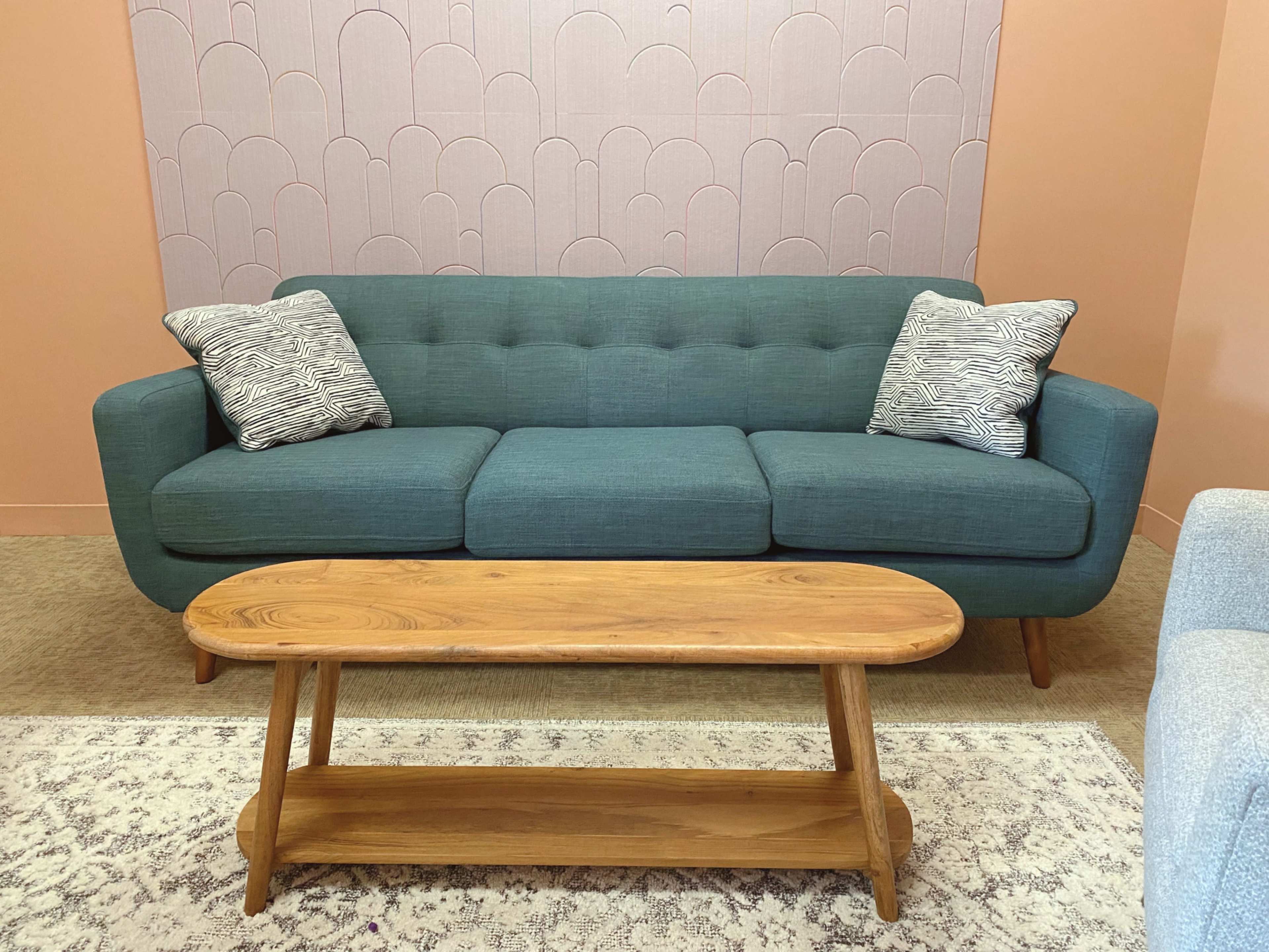 A teal upholstered sofa with two patterned pillows rests in front of a wooden coffee table on a patterned rug.