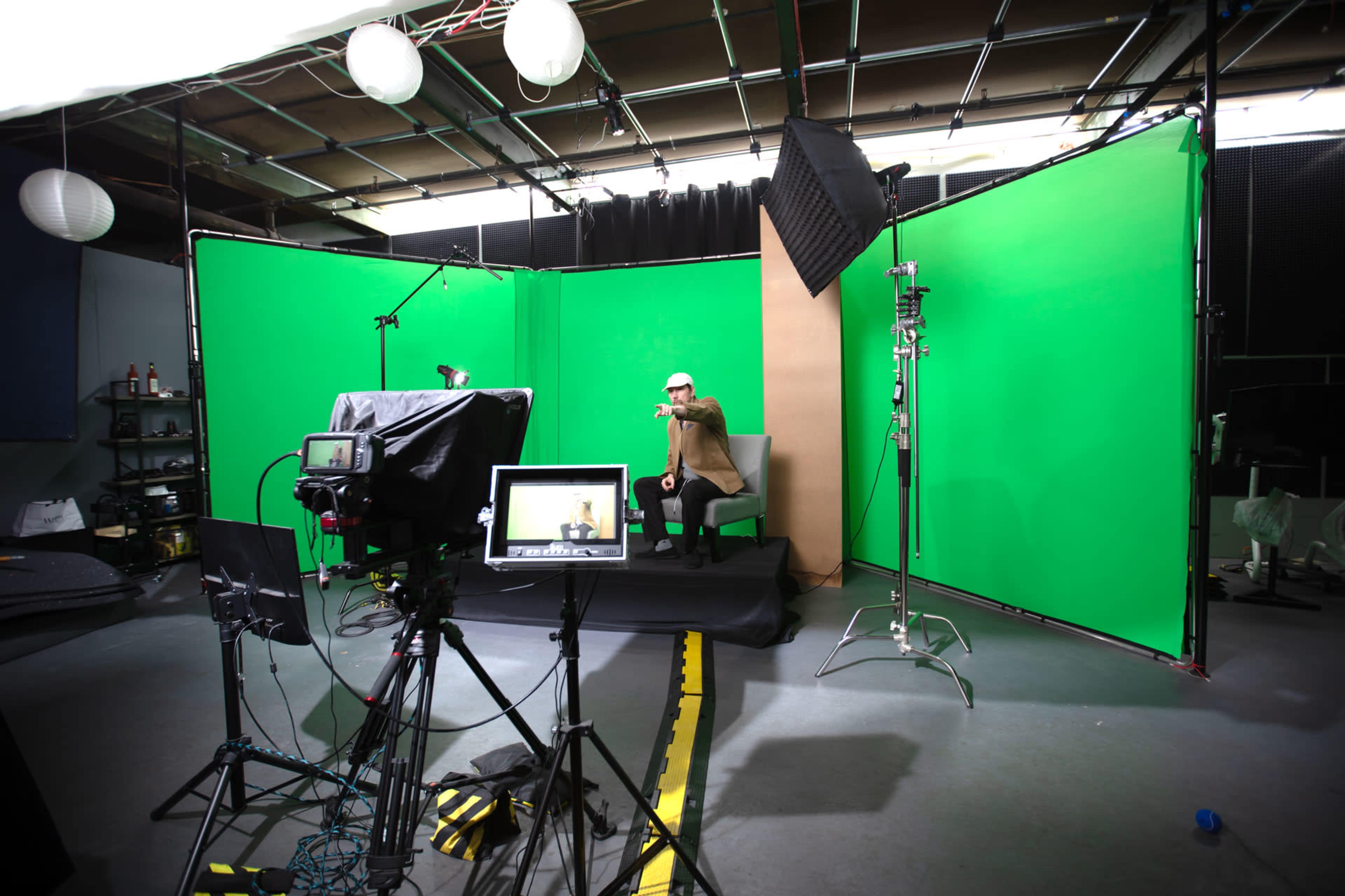 A man sits in a chair in front of green screens and various filming equipment in a studio setting.