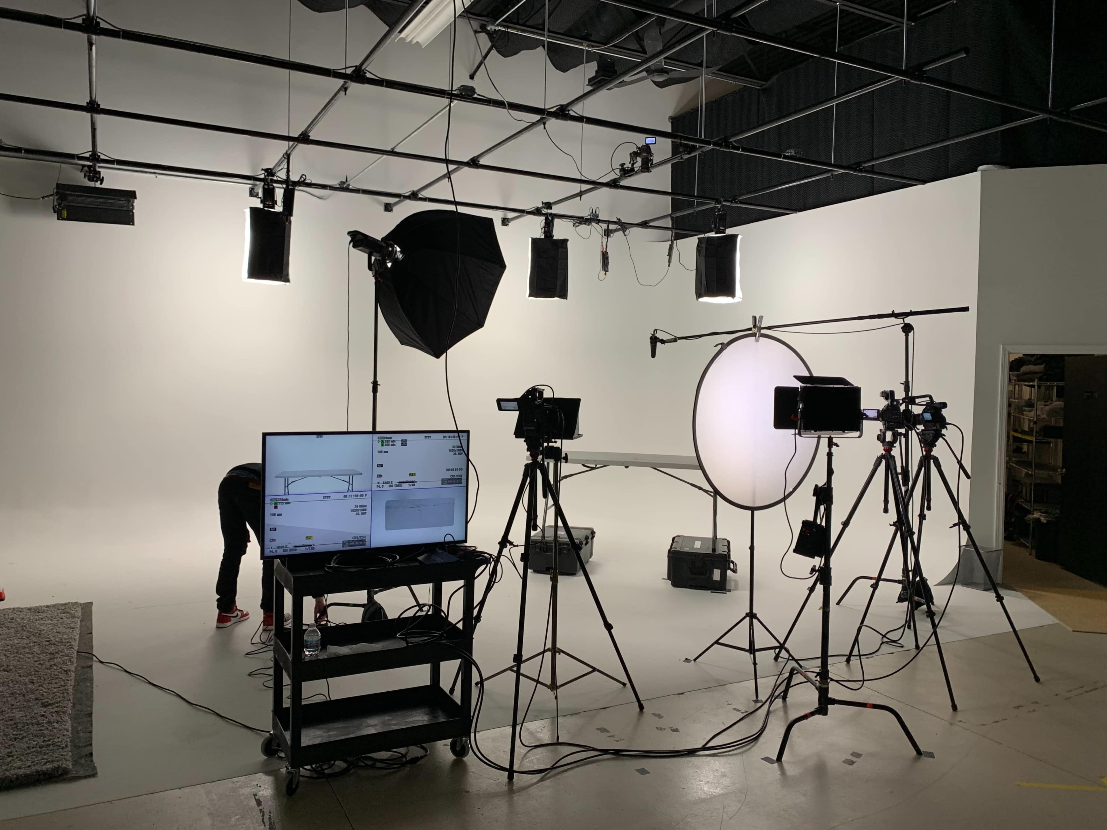 A photography studio is equipped with various lighting setups, cameras, and a monitor on a cart, all arranged in front of a plain white backdrop.
