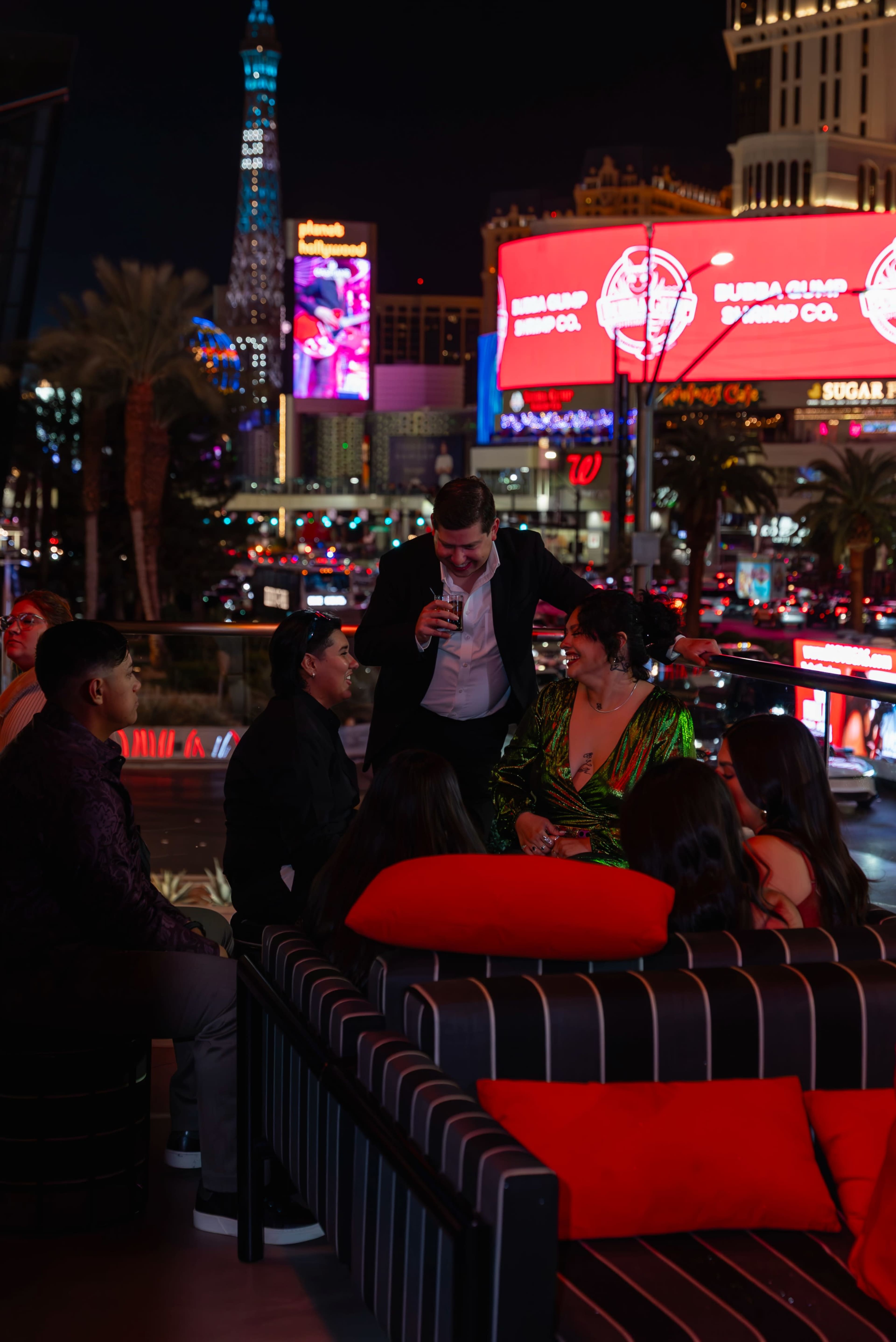A group of people socializes on a rooftop lounge with colorful neon lights and a bustling cityscape in the background.