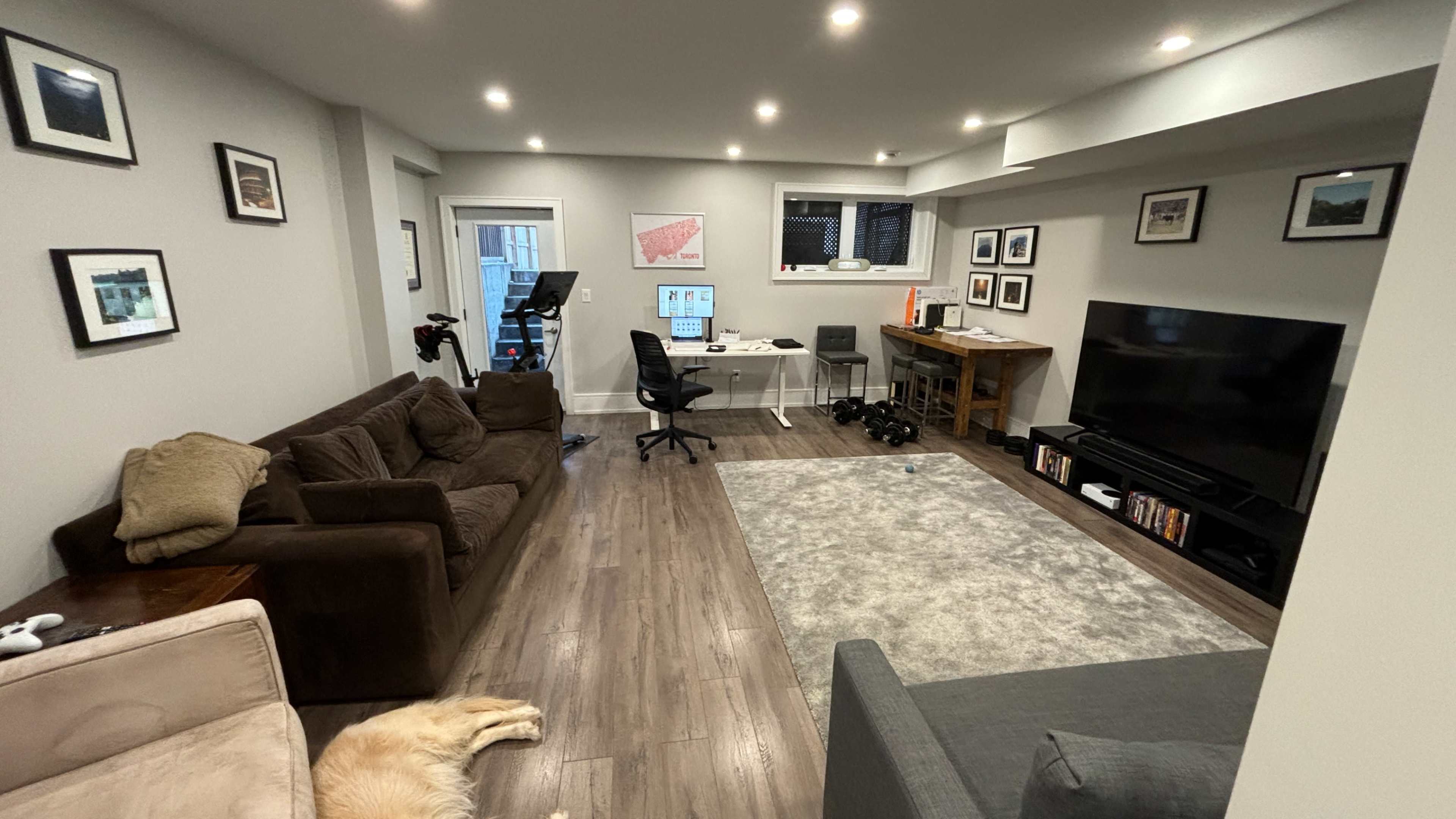 A modern living room featuring two couches, a TV, a desk with computers, and a light-colored rug on hardwood flooring.