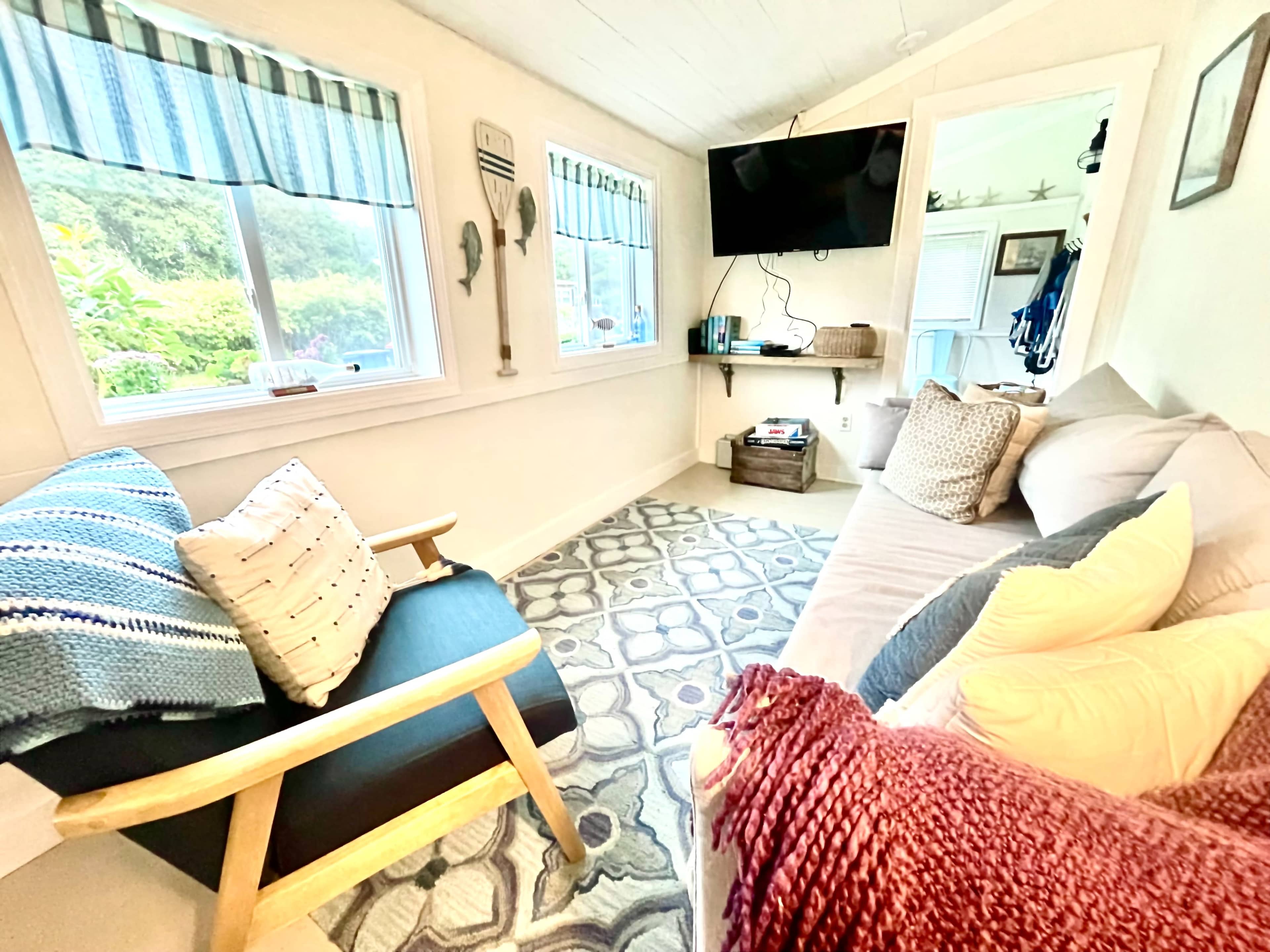 A cozy room with a sofa, an armchair, a television mounted on the wall, and large windows letting in natural light.
