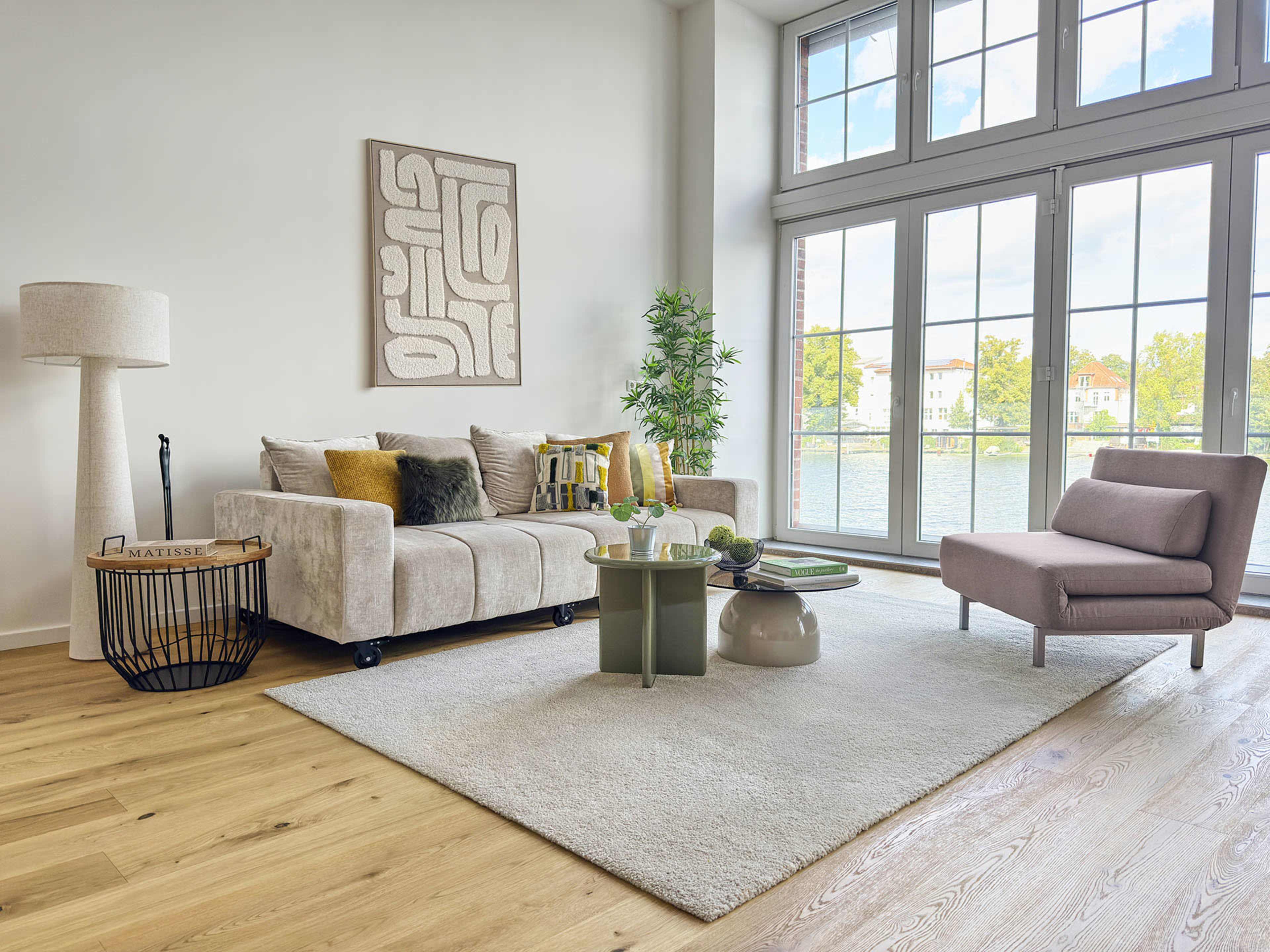 The living room features a light-colored sofa, a modern chair, a round coffee table, and large windows overlooking a body of water.
