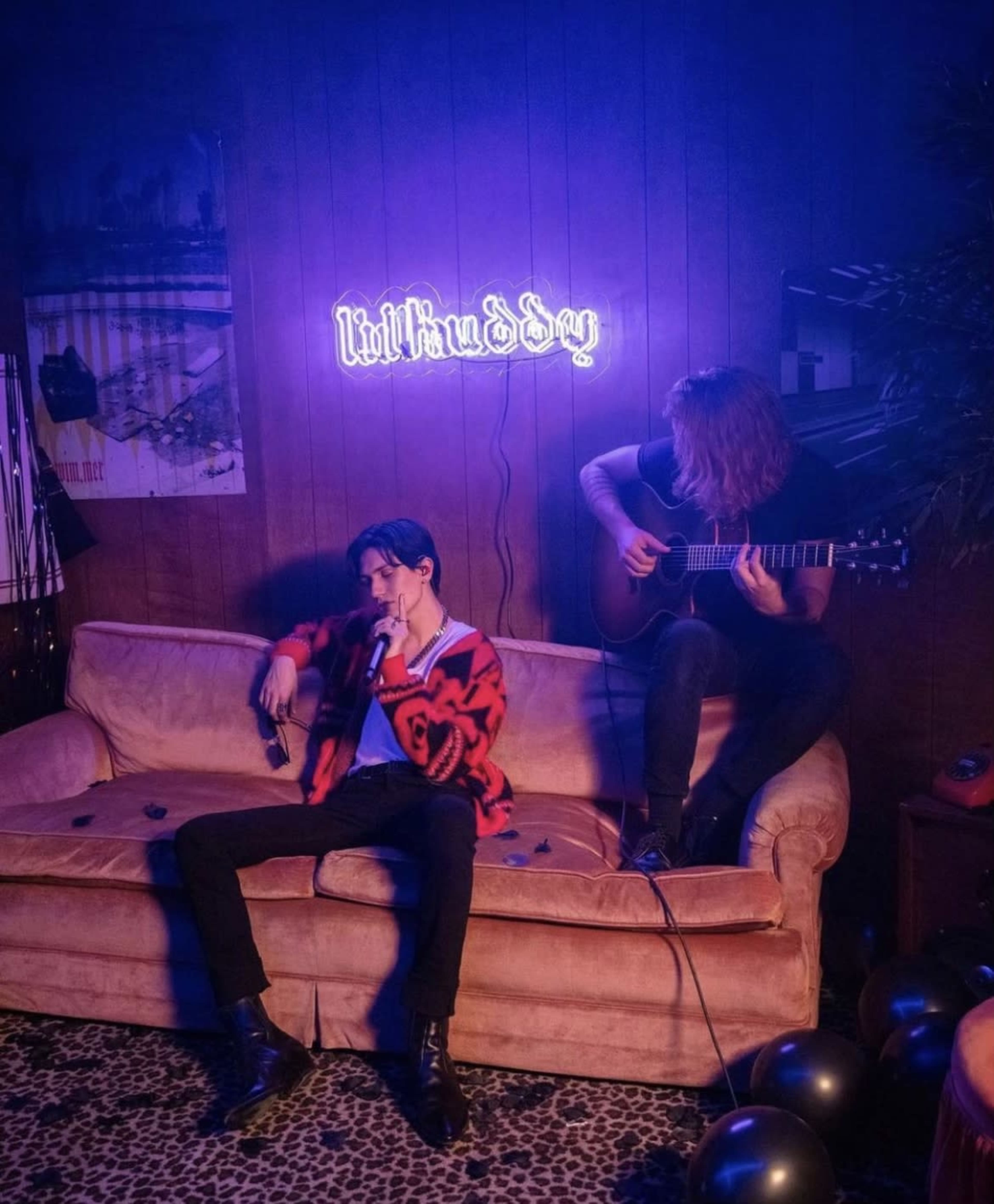 A musician plays guitar while another sits on a couch in a dimly lit room with a neon sign.