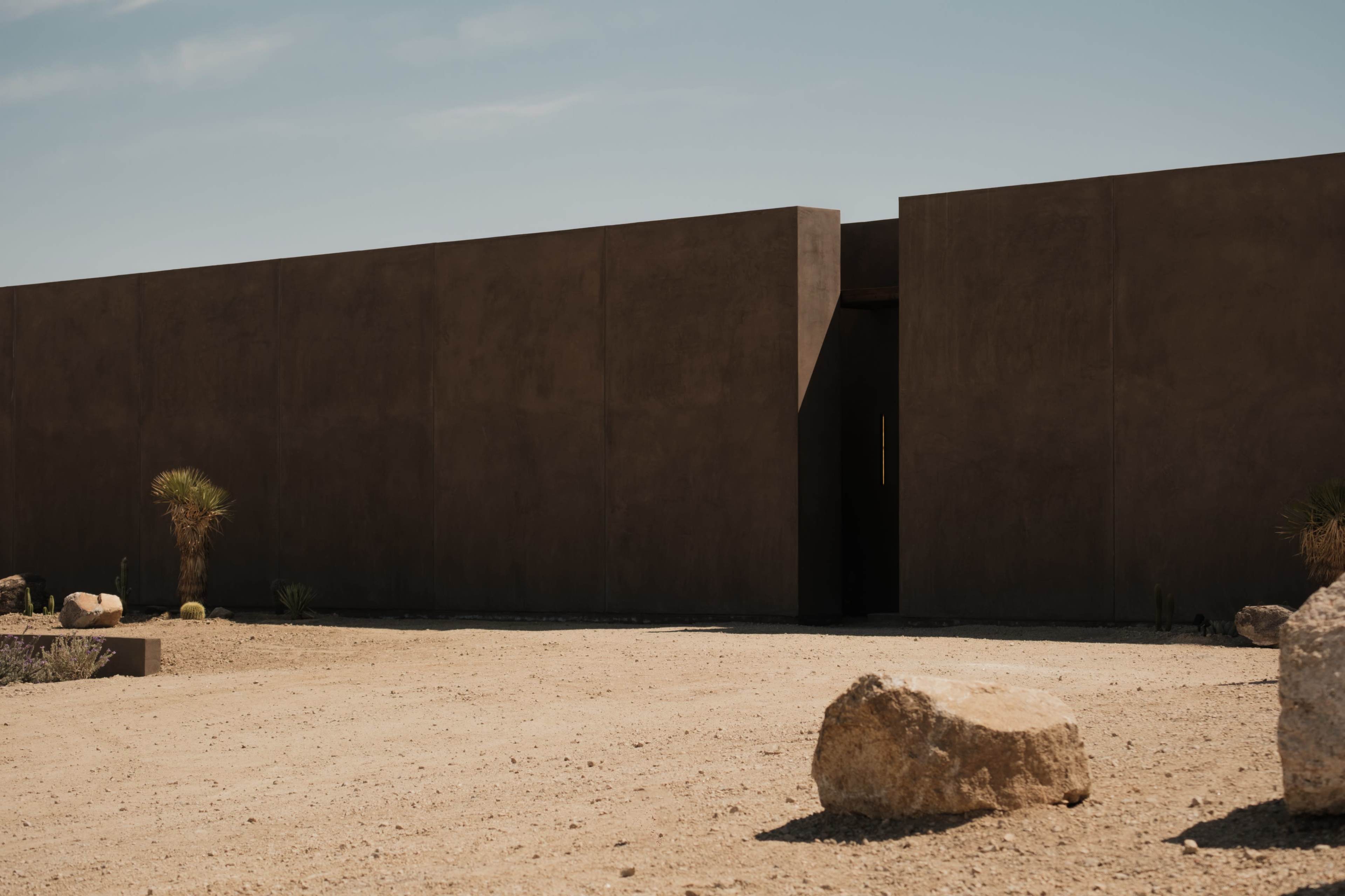 The image shows a modern, minimalist building made of rough-hewn concrete set against a clear blue sky in a barren landscape.