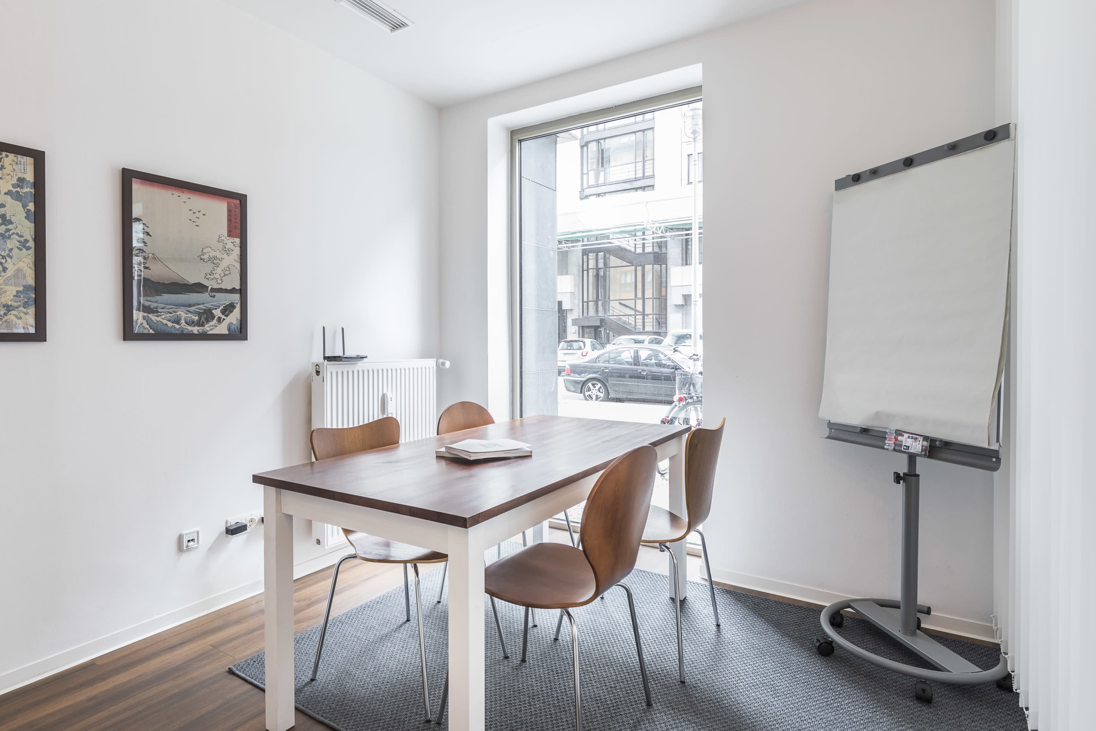 A small conference room features a wooden table with four chairs, a flip chart, and a window overlooking a street.