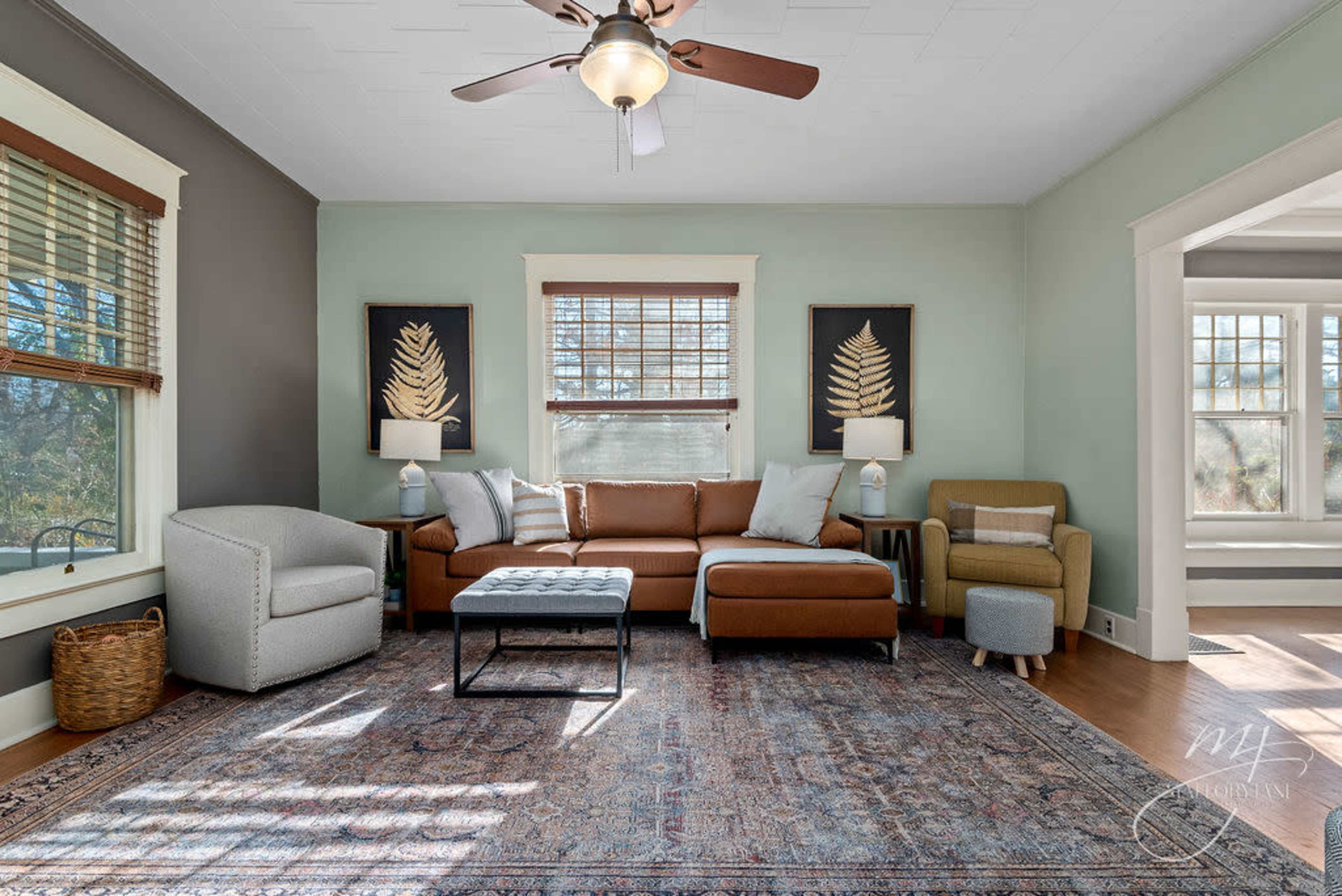 The image shows a living room featuring a brown sectional sofa, a gray armchair, and a patterned area rug, with wall art and large windows providing natural light.