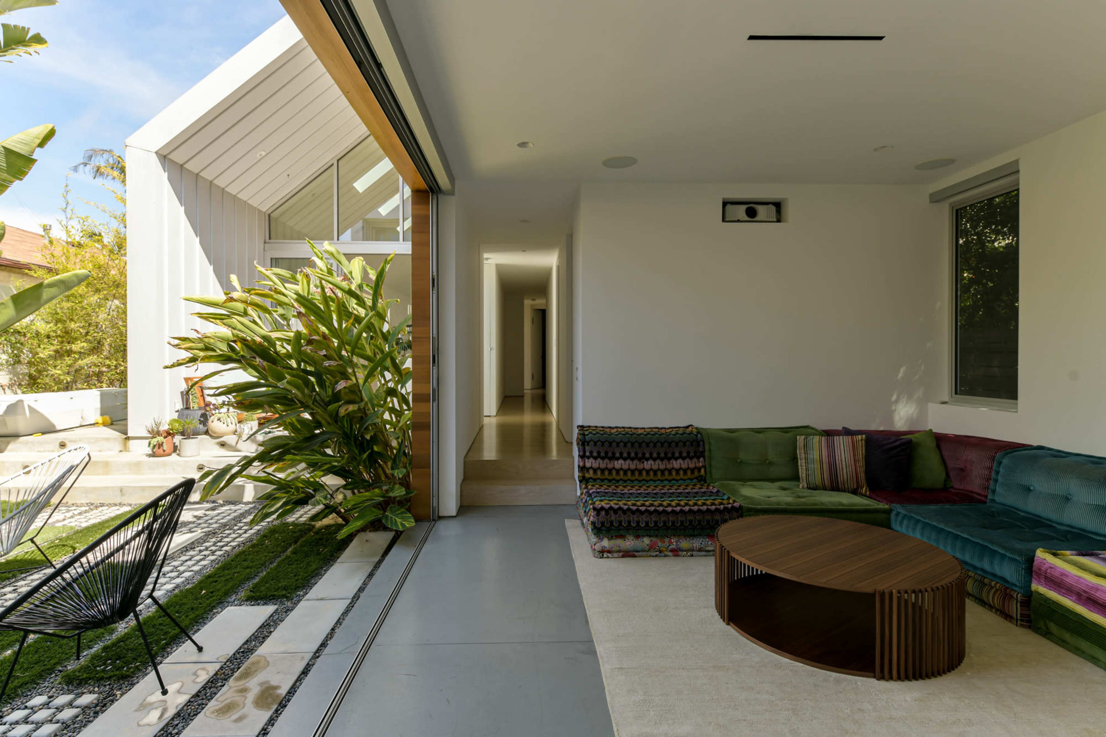 A modern living space with a green and multicolored sofa, a round wooden coffee table, and large windows opening to a garden area lined with plants.