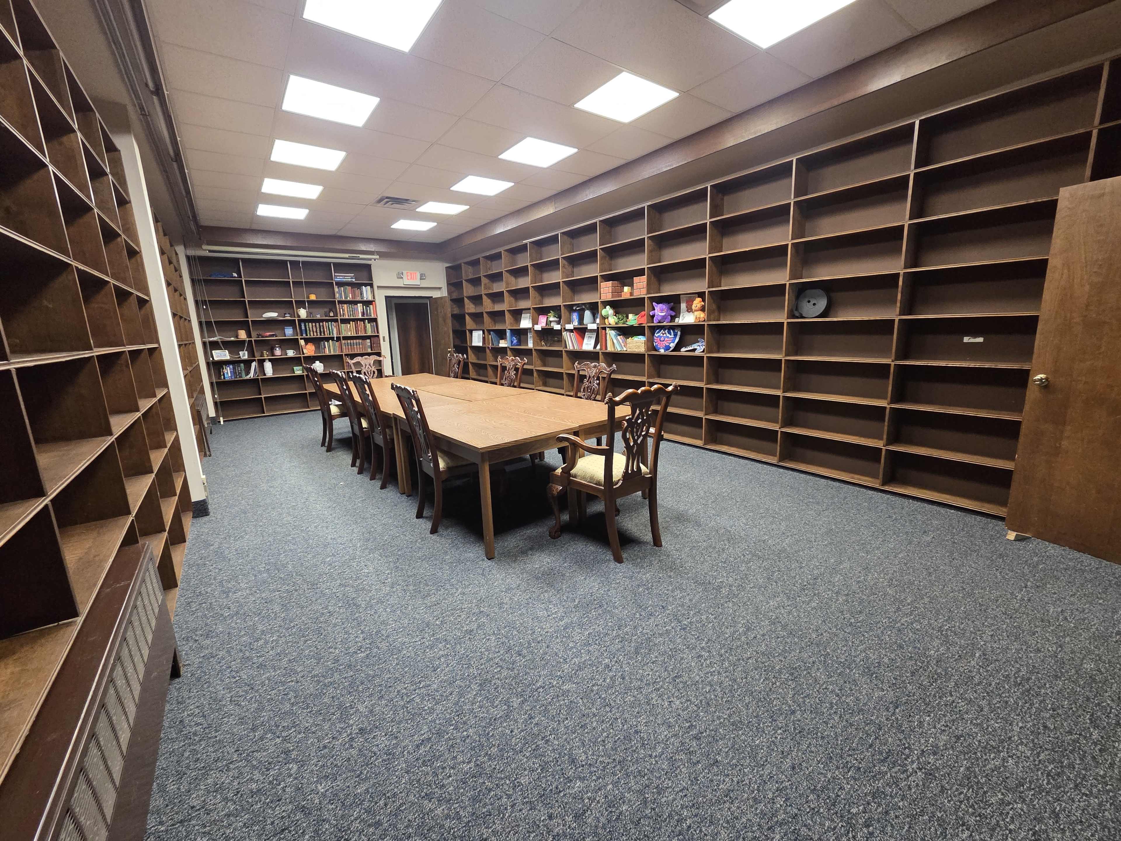The image shows a large meeting room with a long wooden table surrounded by chairs, set against a backdrop of empty wooden bookshelves.