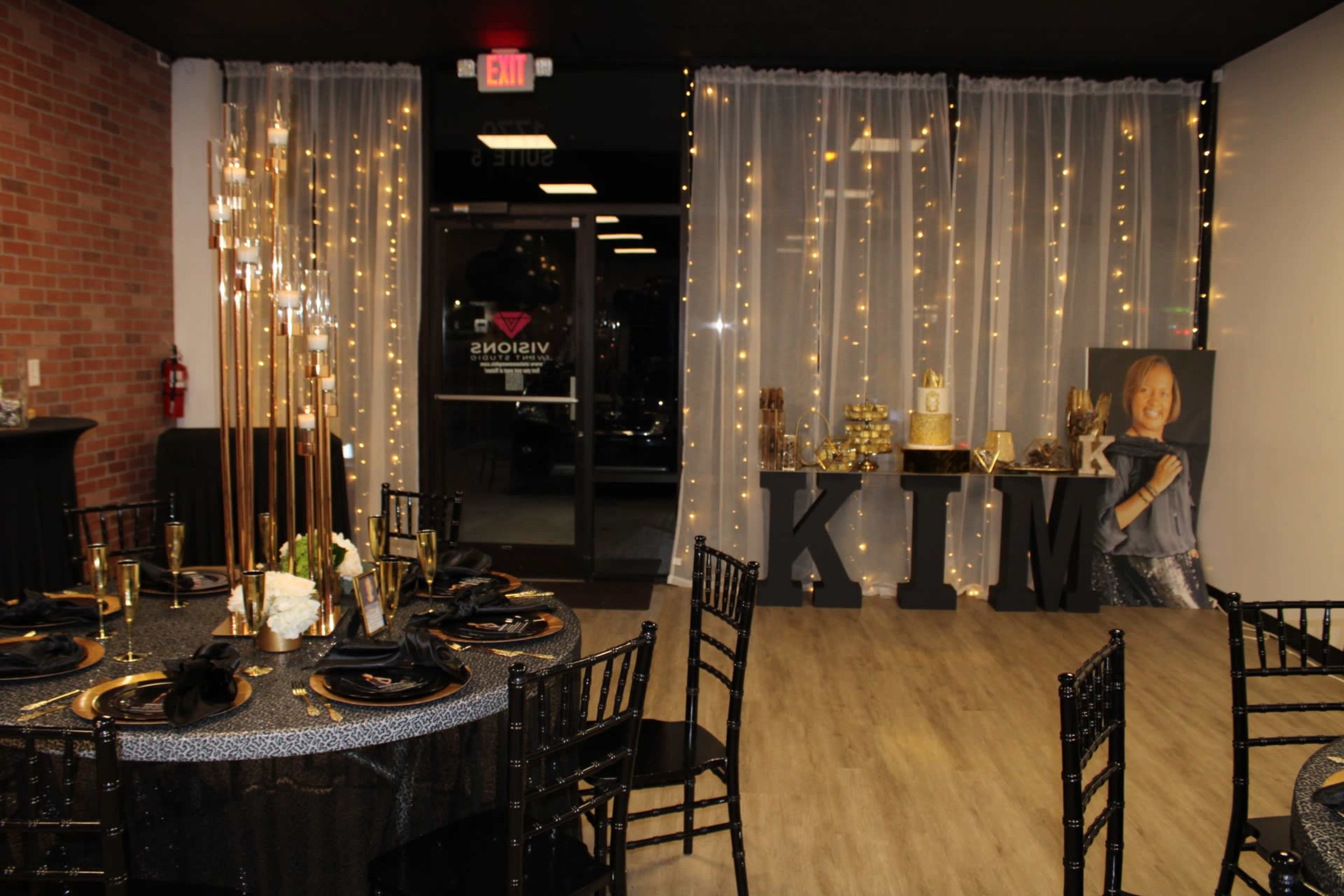 A decorated event space with black tables, gold accents, and a backdrop featuring a large "KIM" display along with a framed portrait.