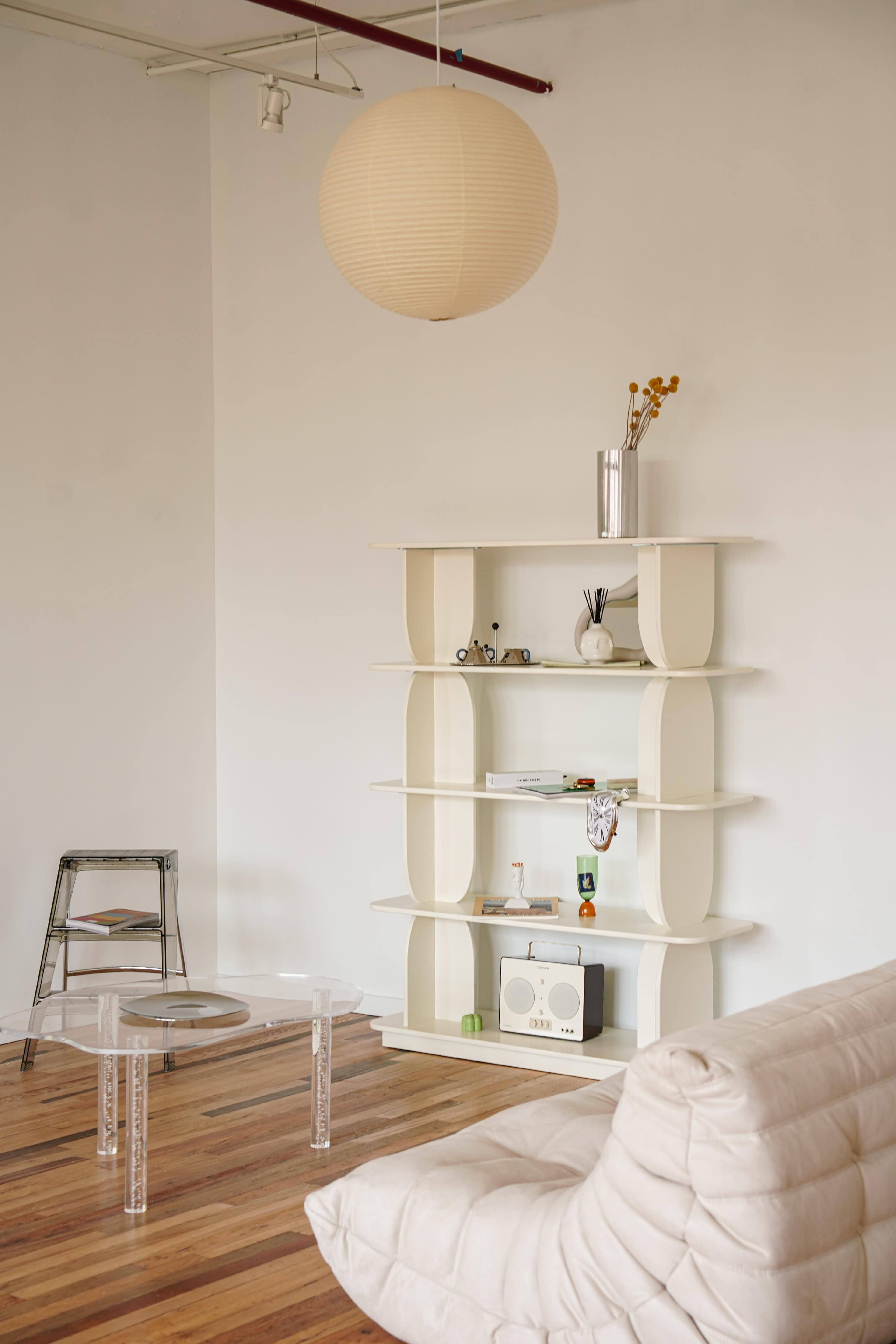 A minimalist living space featuring a transparent coffee table, a white shelf with various decorative items, and a light-colored sofa.