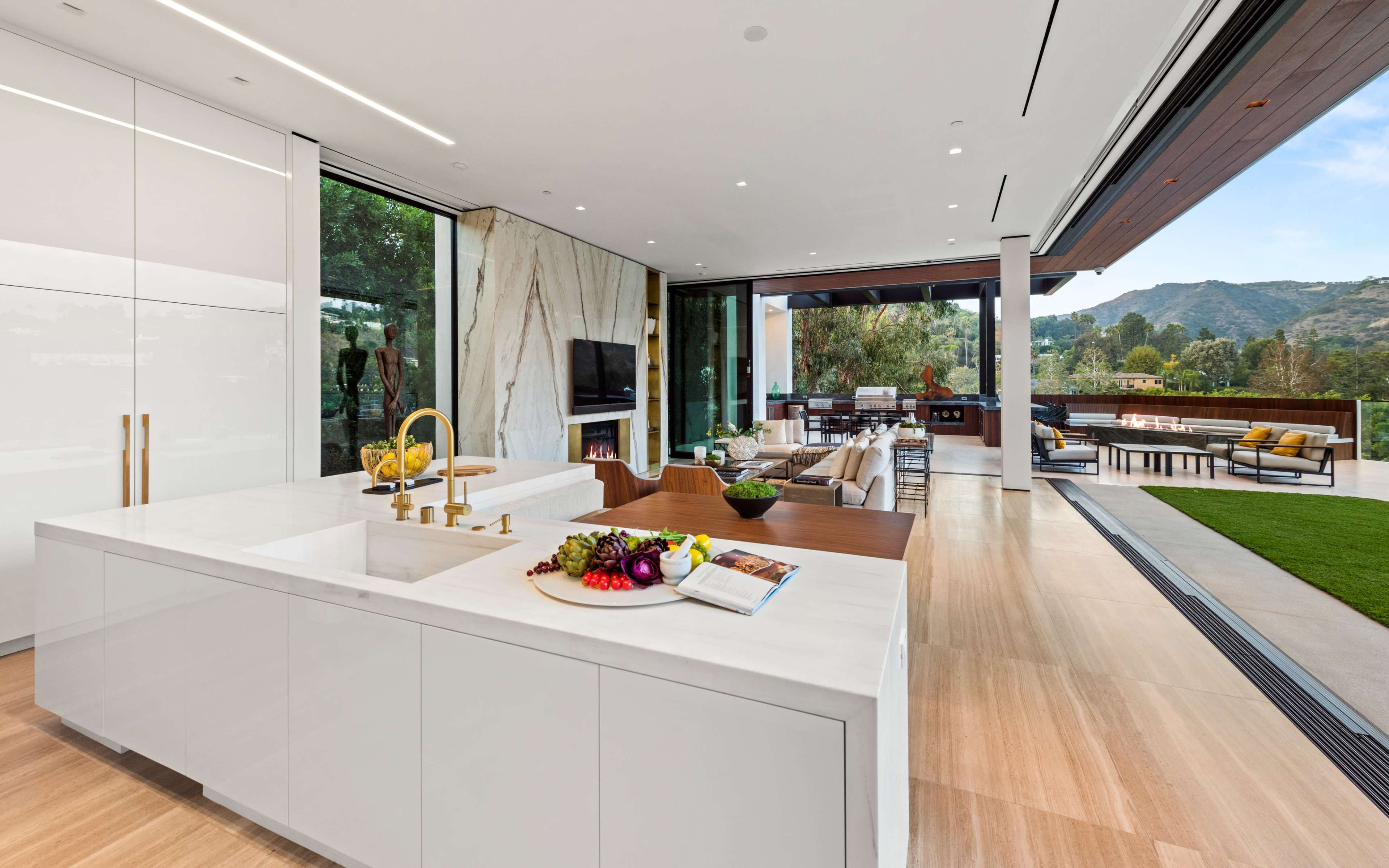 The image shows a modern kitchen with a large island, sleek cabinetry, and an open layout that connects to a living area with floor-to-ceiling windows showcasing a view of the outdoor patio and mountains.