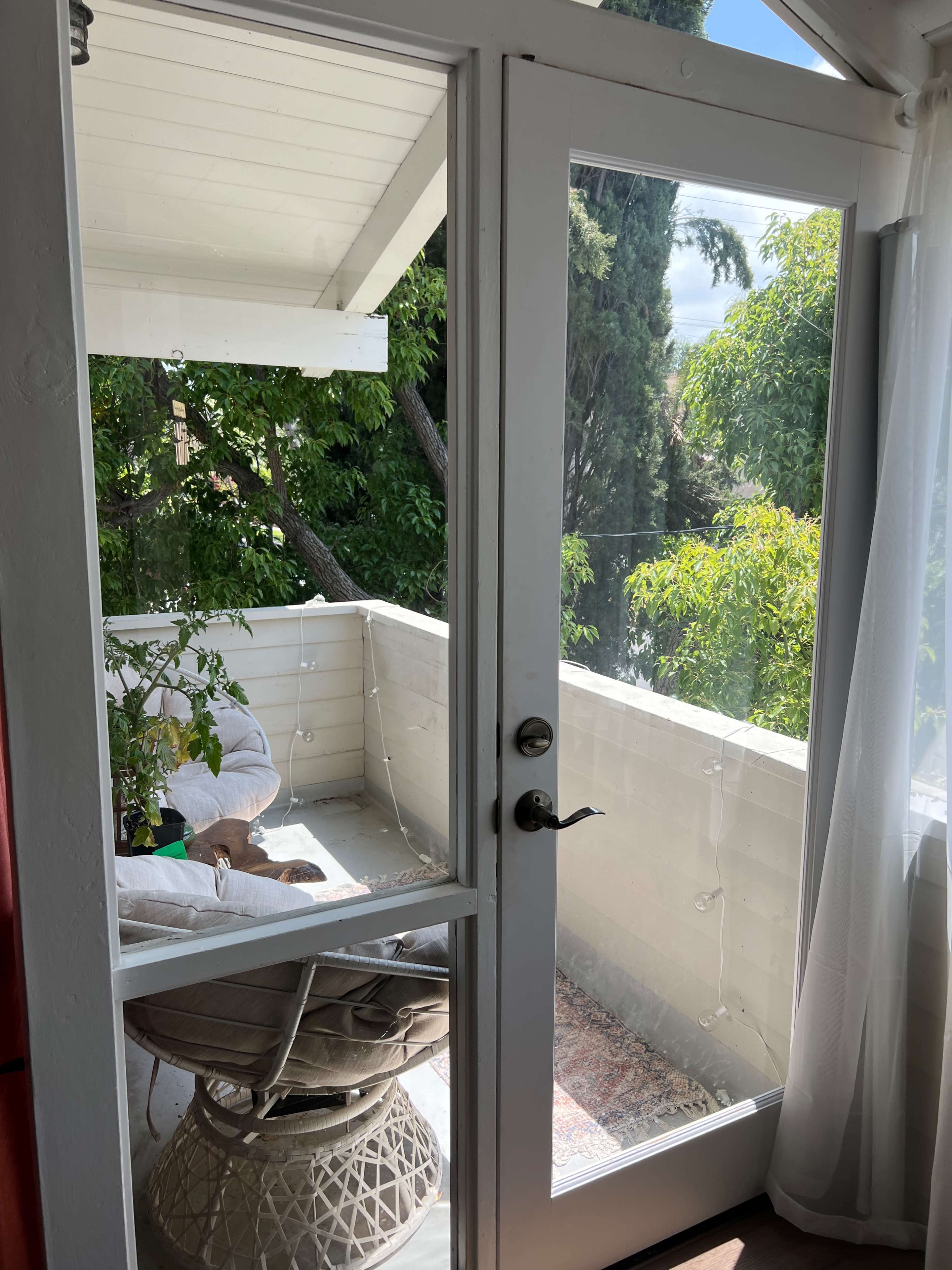 The image shows a balcony view through a glass door, featuring a chair, plants, and greenery outside.