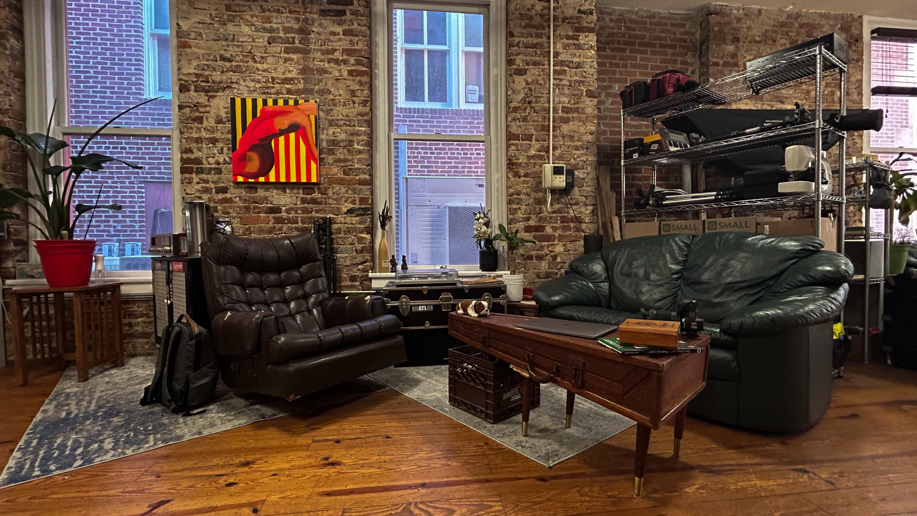 A cozy living space with a brown leather armchair and a green couch on a wooden floor, surrounded by exposed brick walls and large windows.