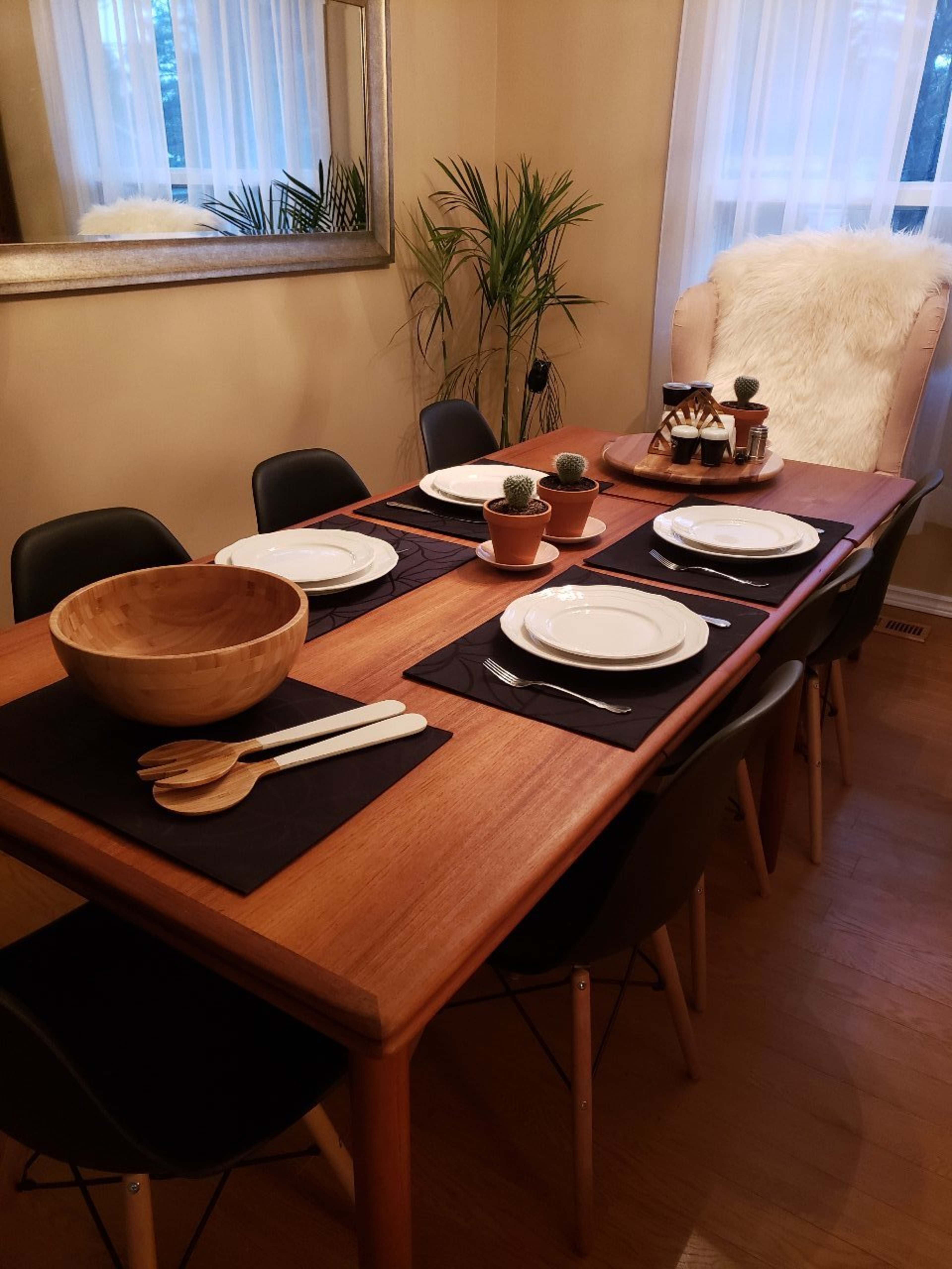 A wooden dining table is set with white plates and utensils, surrounded by black chairs, with a bowl and potted plants as centerpieces.
