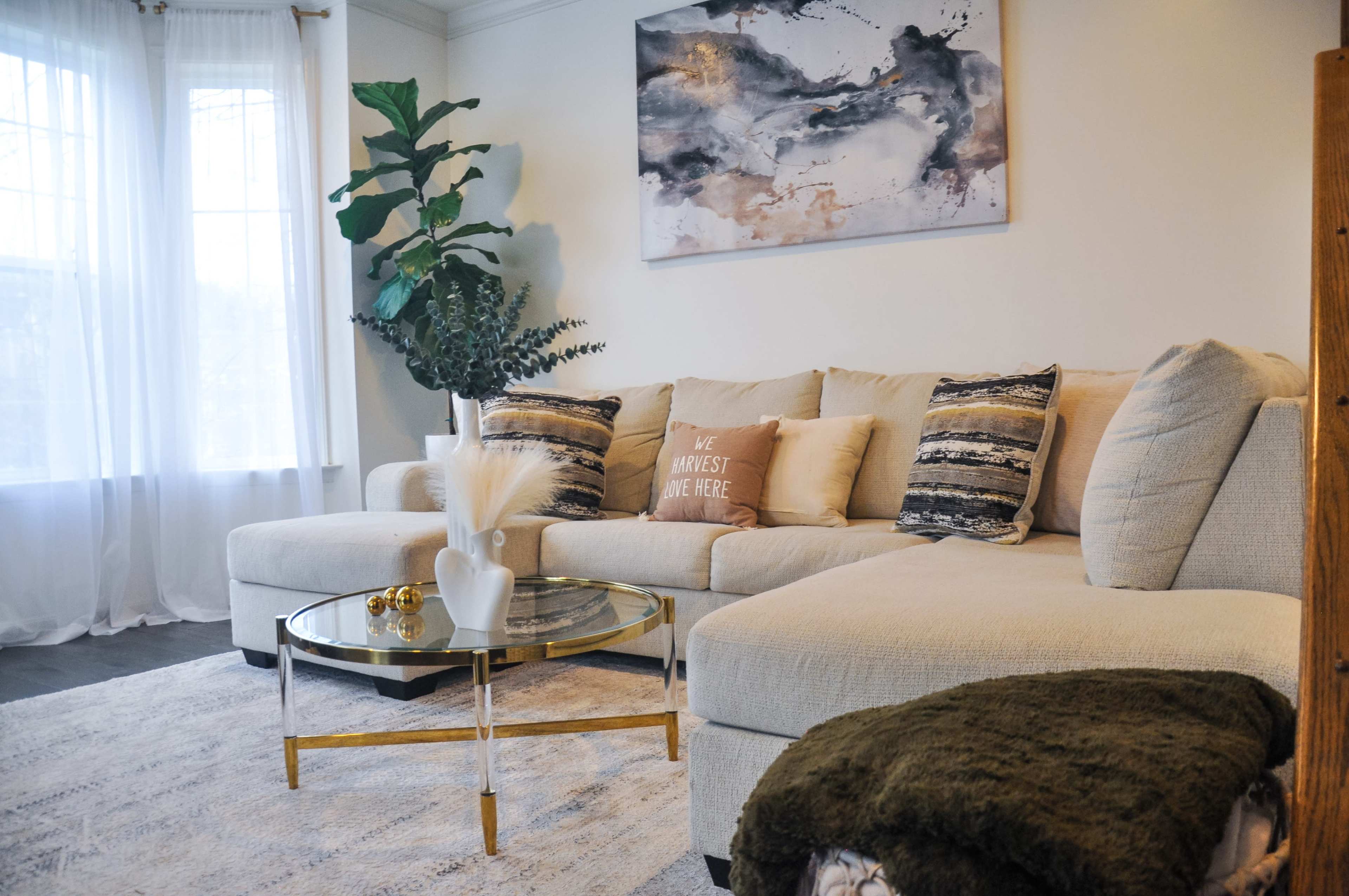 The image shows a cozy living room featuring a beige sectional sofa with decorative pillows, a round glass coffee table, and a large abstract artwork on the wall above.